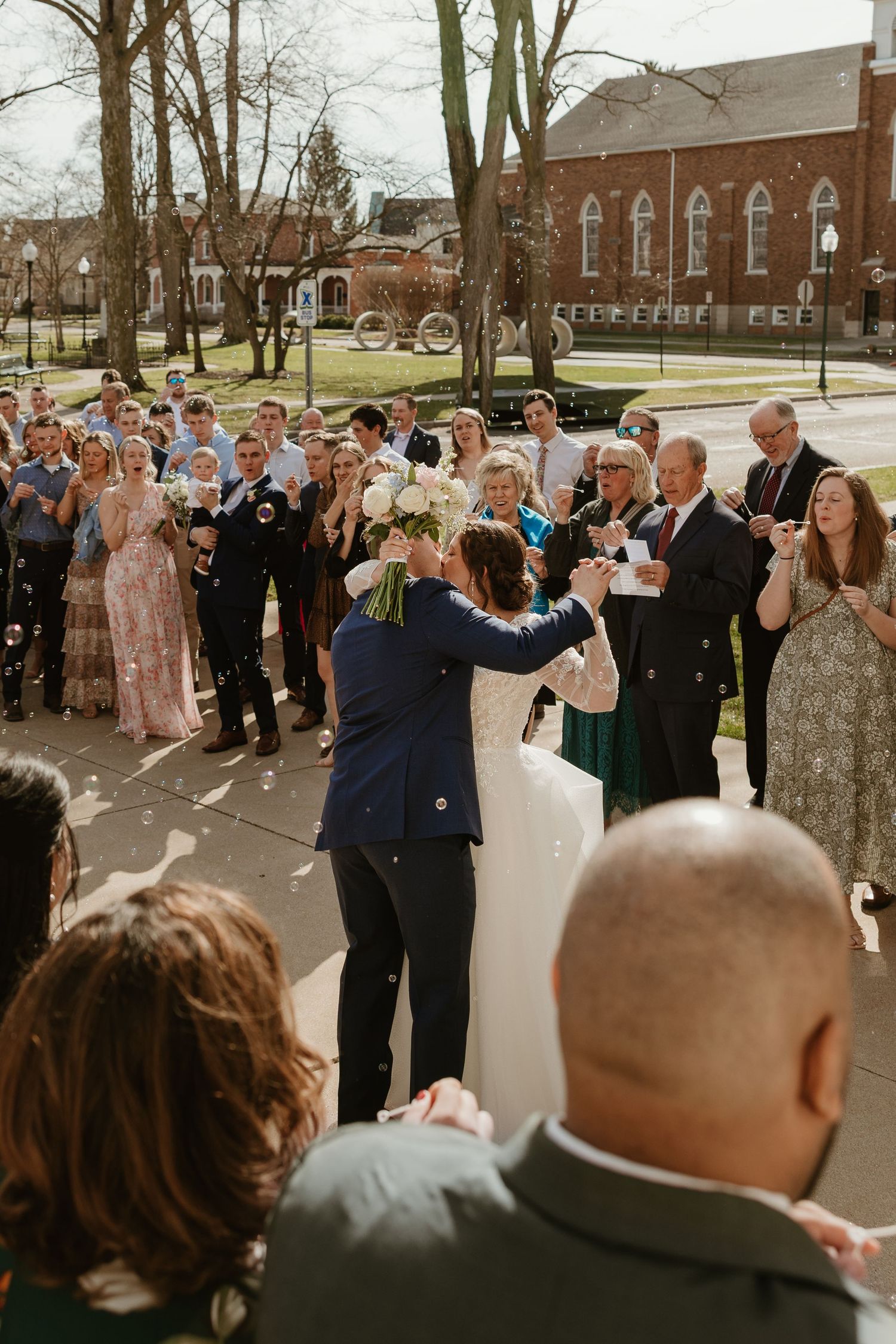 Newlywed couple kisses outside church surrounded by cheering guests blowing bubbles after wedding ceremony.