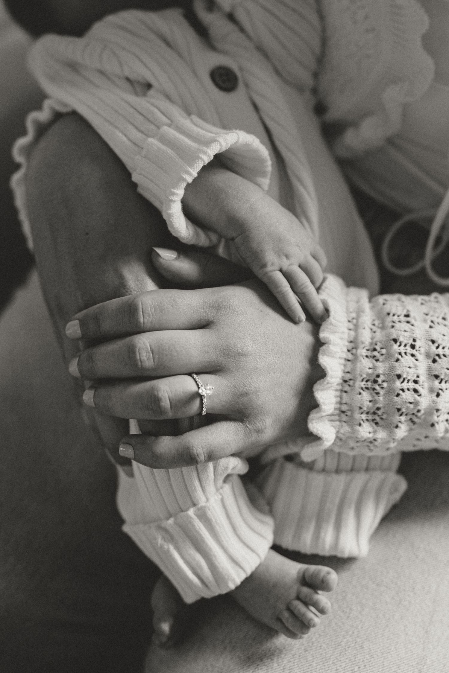 Black and white photo of a mother's hands with a ring cradling her newborn baby's tiny feet and legs.