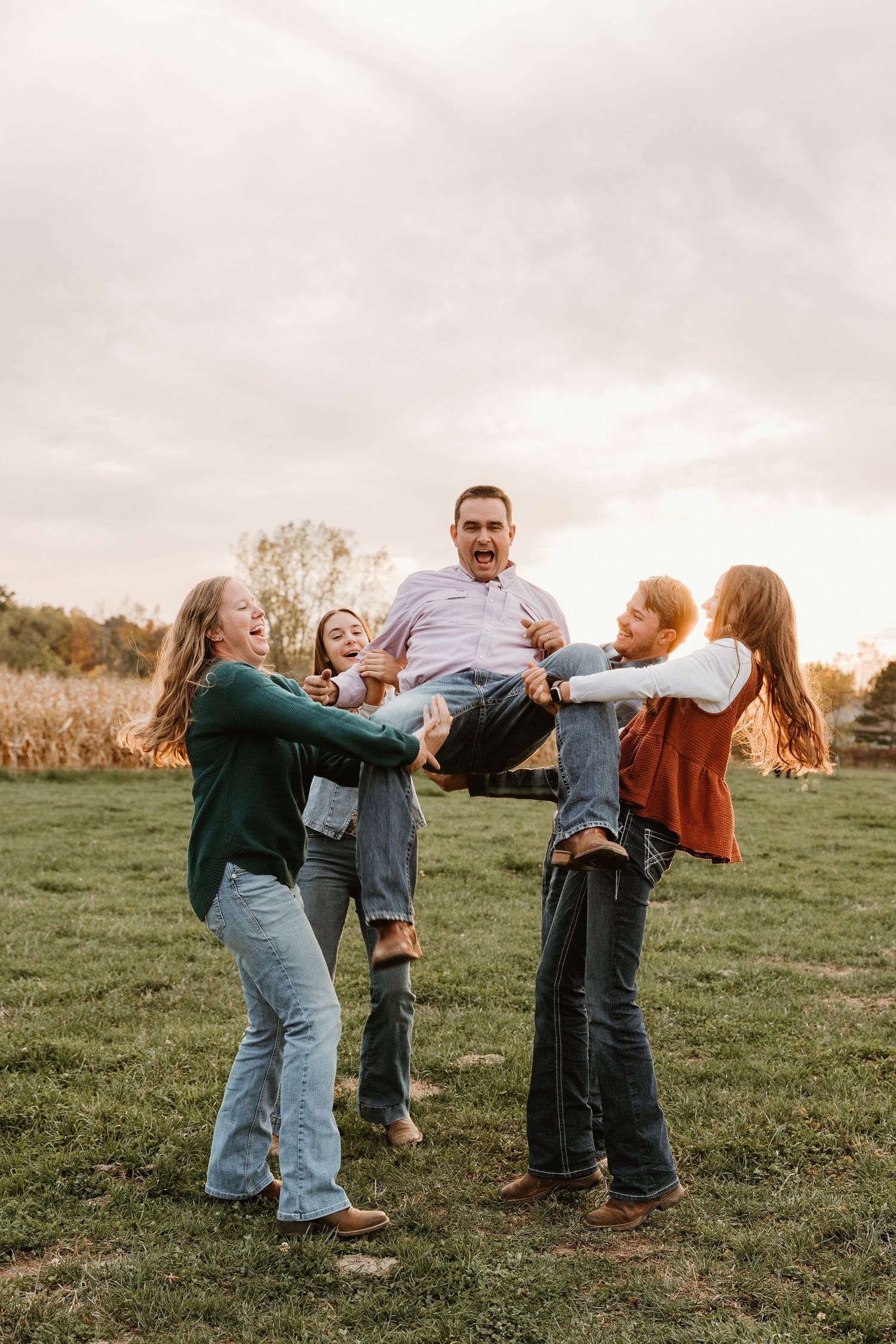 Family laughing outdoors as children lift dad in a fun autumn field photo session.