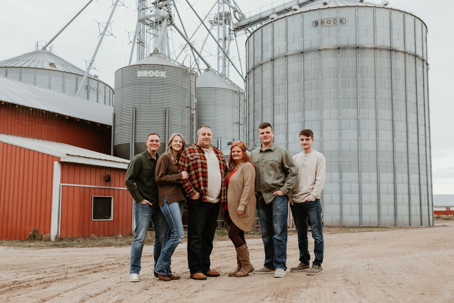 Family of six poses together in front of large grain silos and red barn on a farm.