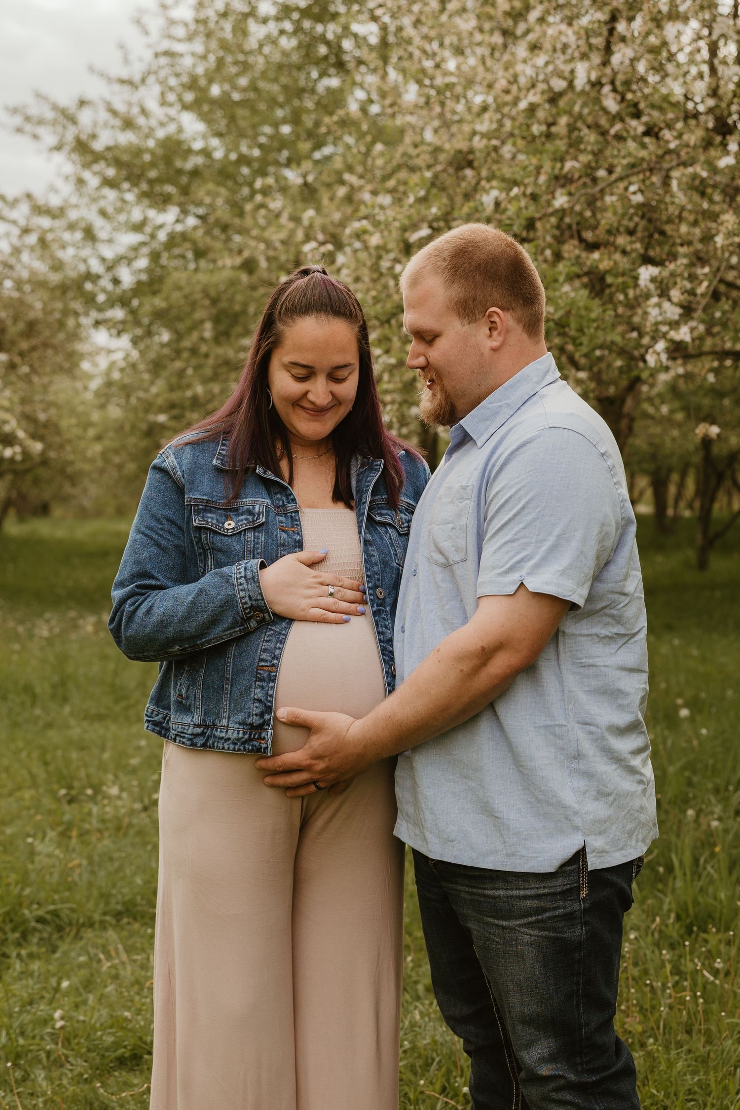 Pregnant couple smiling and cradling baby bump outdoors among blooming apple trees in spring.
