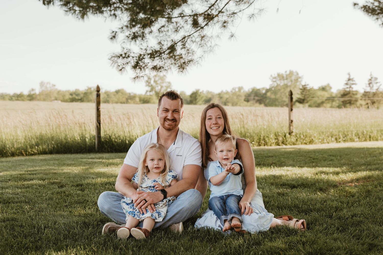 Happy family of four sitting on green grass outdoors, parents holding two toddlers, smiling in warm golden sunlight.