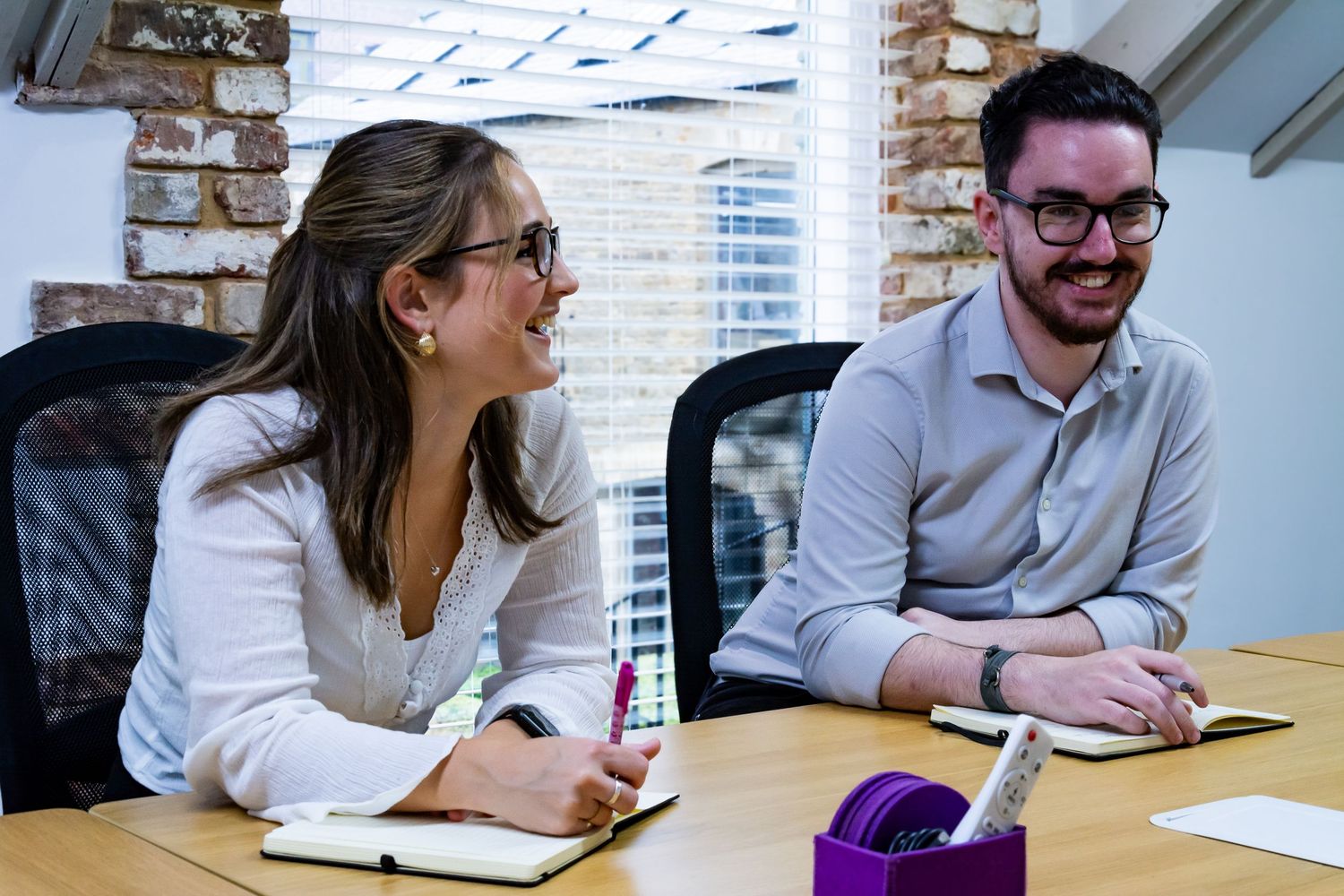 Two young professionals smiling and taking notes during a collaborative office meeting.