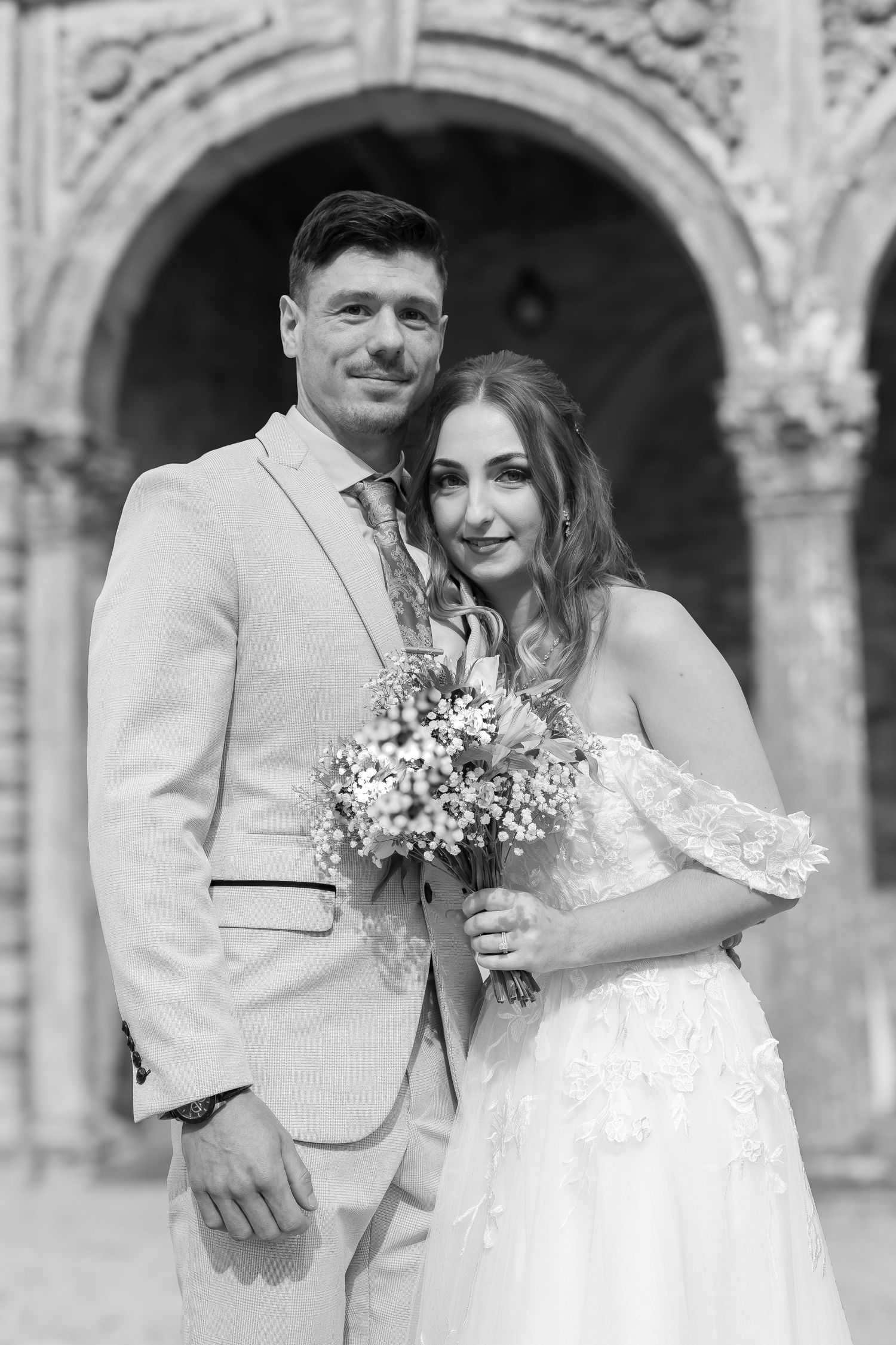 Black and white portrait of smiling bride and groom embracing before a stone Gothic archway.