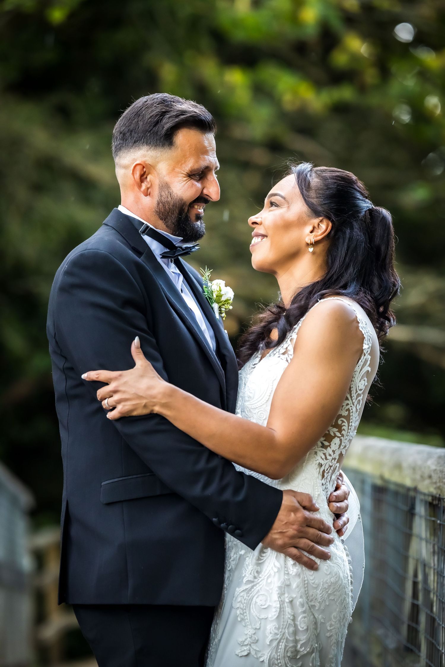 Bride and groom share romantic embrace outdoors, groom in black tuxedo and bride in lace wedding dress.