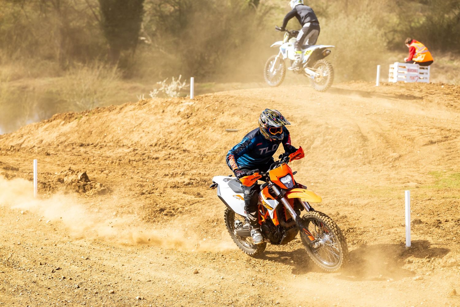 Motocross riders racing on a dusty dirt track, with the lead rider on an orange KTM bike kicking up clouds of dust.