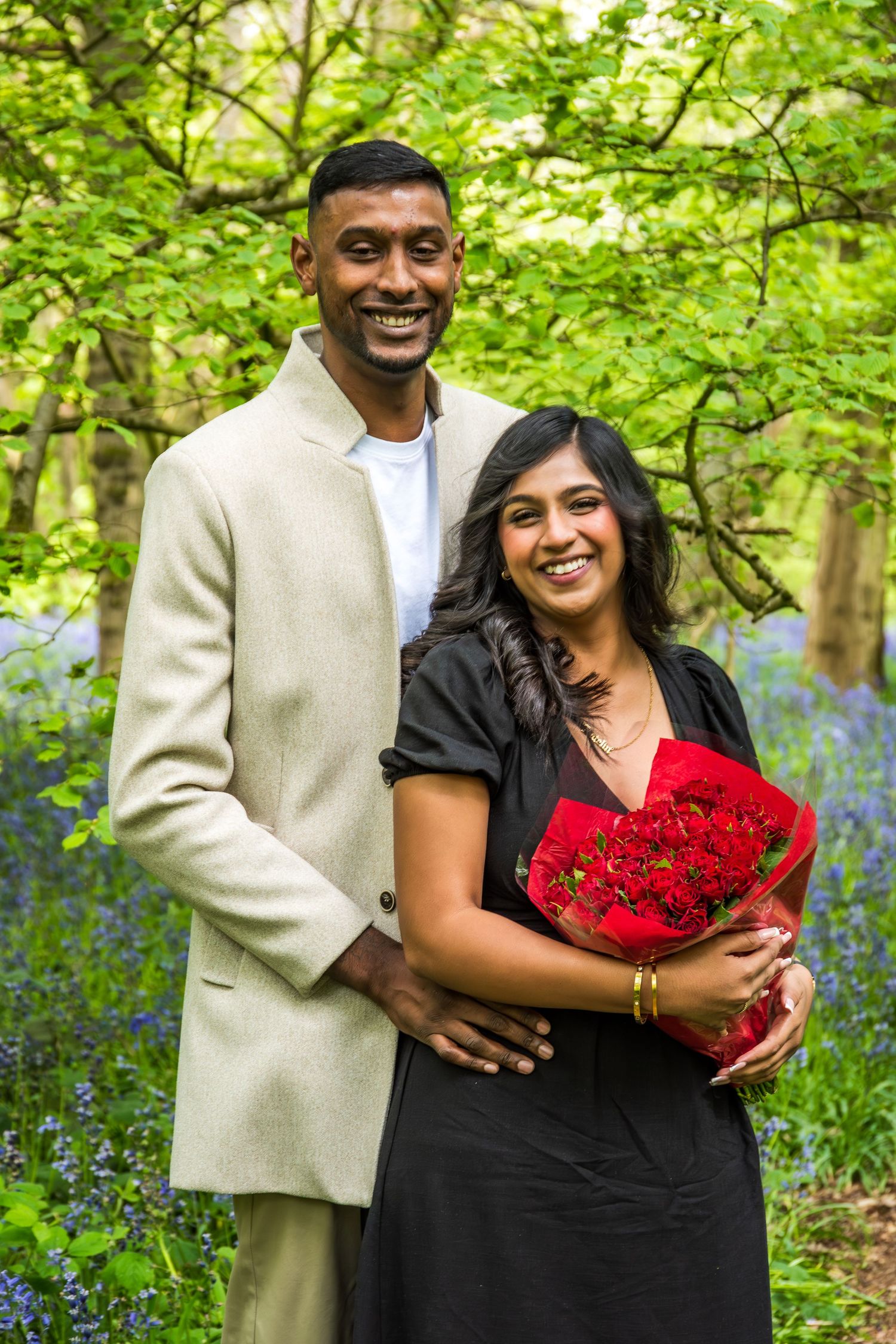 Smiling couple posing outdoors in bluebell woods, woman holding red roses bouquet, man in beige blazer embracing her.