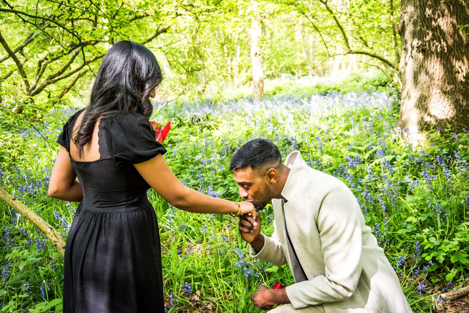 Man in white suit kneeling to kiss woman's hand in a bluebell forest during a romantic outdoor proposal.