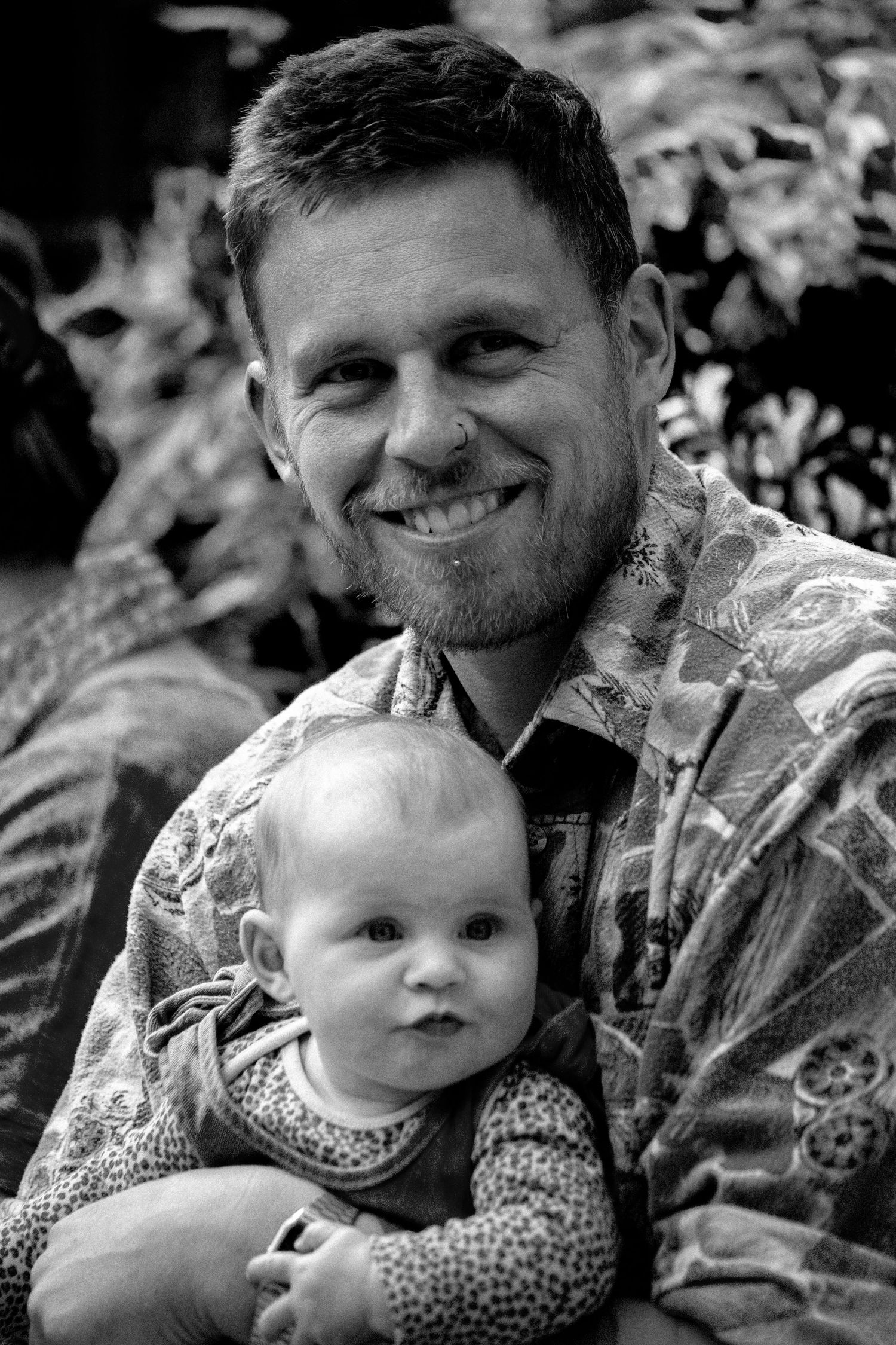 Black and white portrait of smiling father holding curious baby in patterned jacket outdoors.