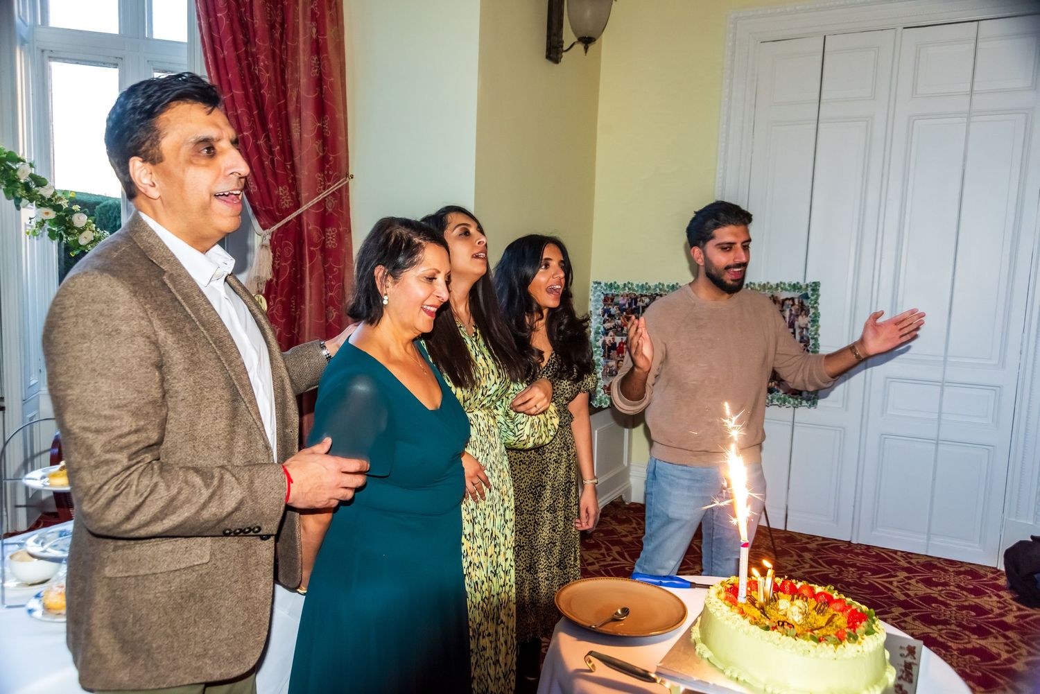 Family gathered around birthday cake with sparklers, celebrating indoors in festive decorated room.