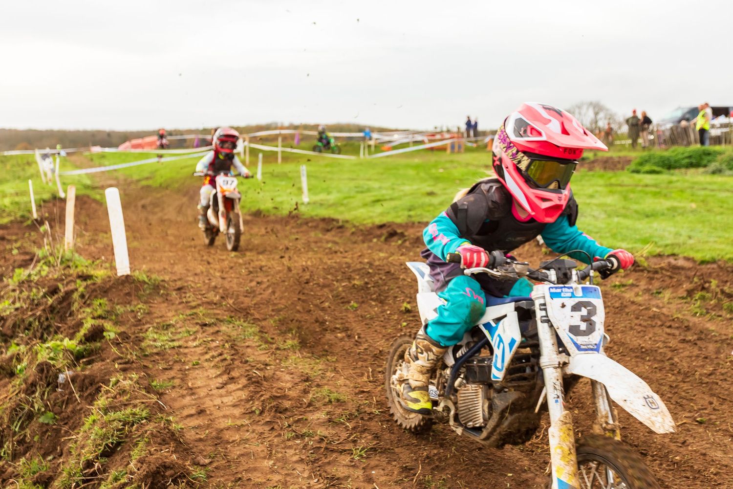 Young motocross riders race on a muddy dirt track, with rider number 3 in teal gear leading the competition.