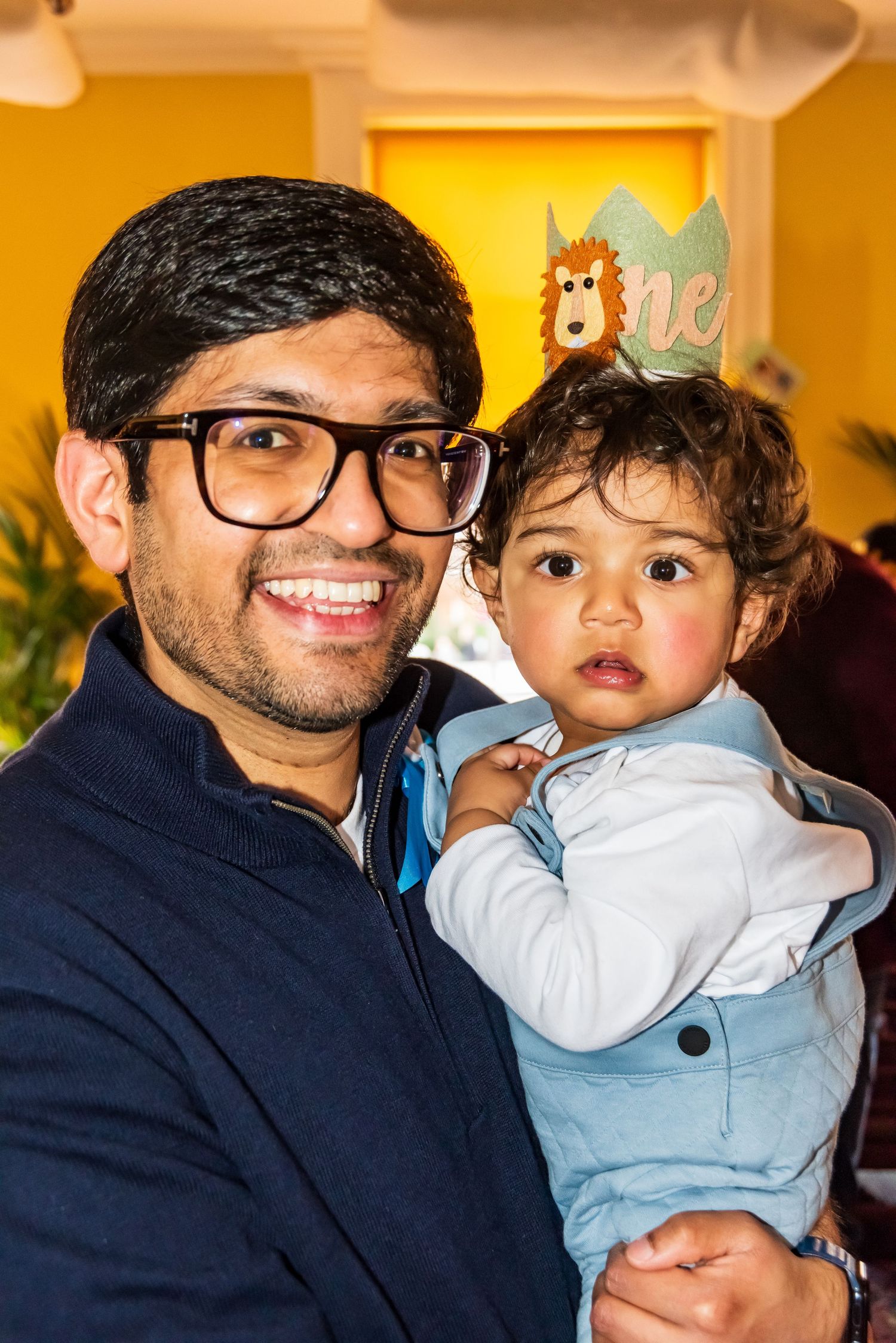 Smiling man with glasses holding a baby at a first birthday party.