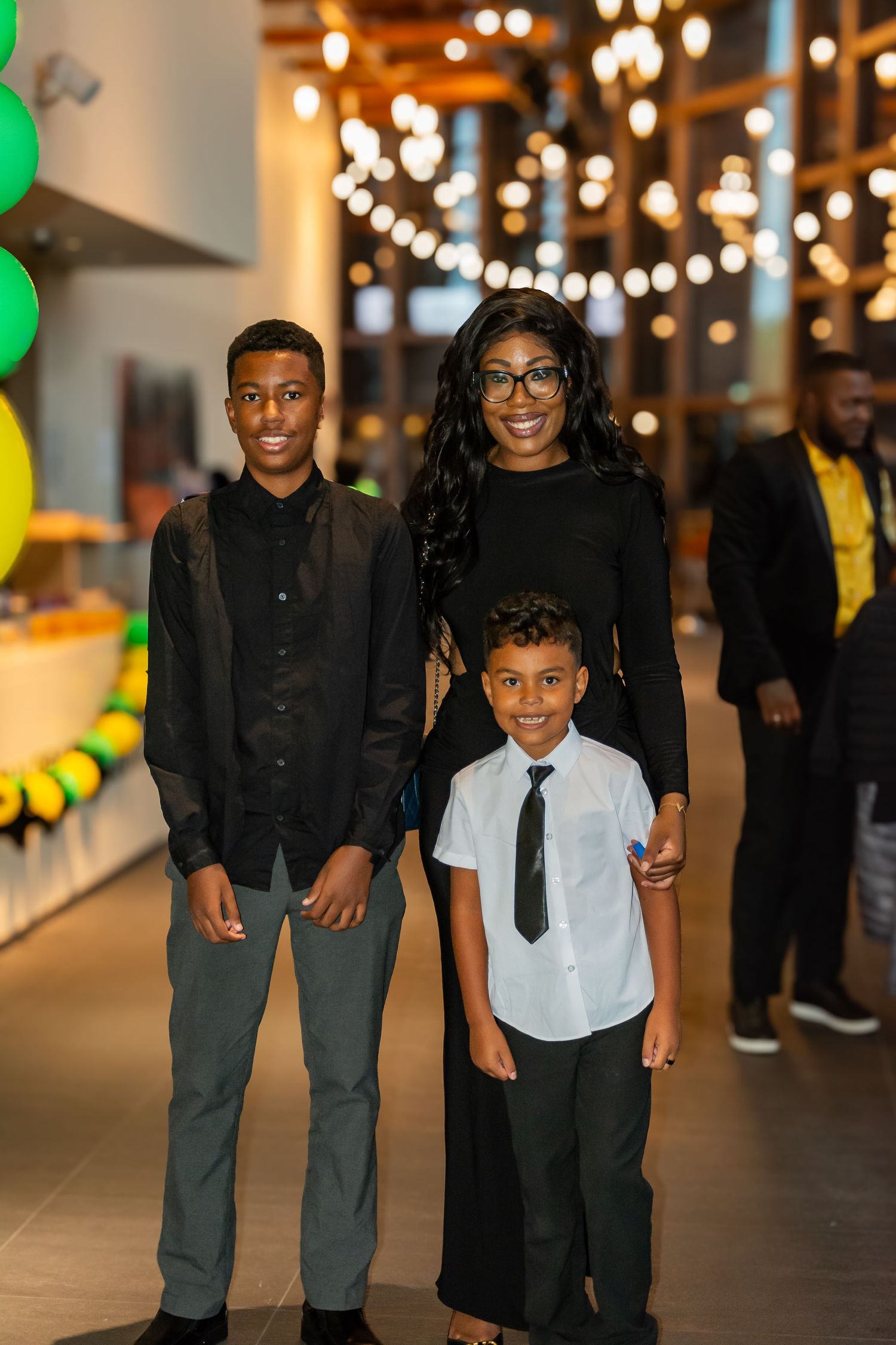 Woman with glasses poses with two boys in formal attire at a festive event with string lights.