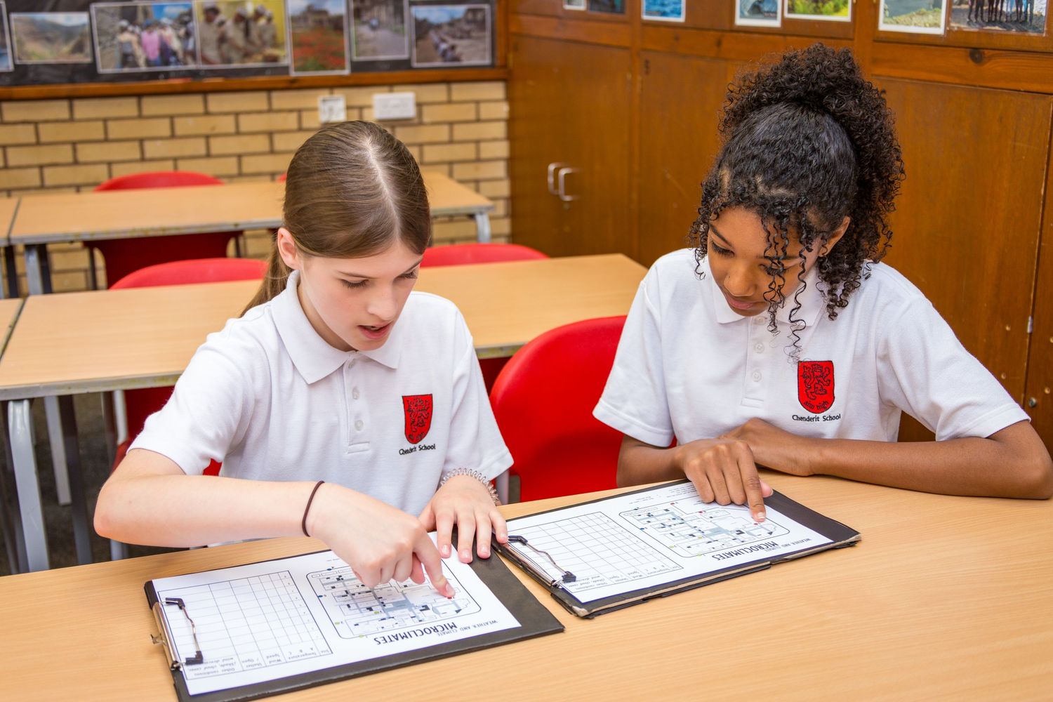 Two school girls in white uniforms collaborate on worksheets at a classroom desk. Photographed by LJPhotostop for school website content.