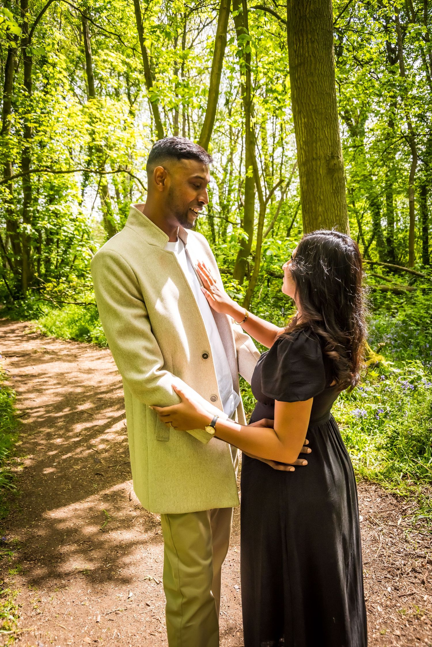 Couple sharing a romantic moment on a sunlit forest path, man in beige suit and woman in black dress.