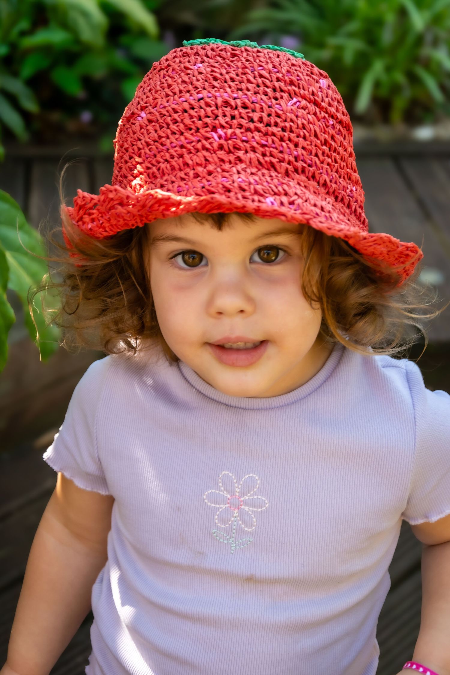 Cute toddler girl with curly hair wearing a red crocheted sun hat and white floral t-shirt outdoors.
