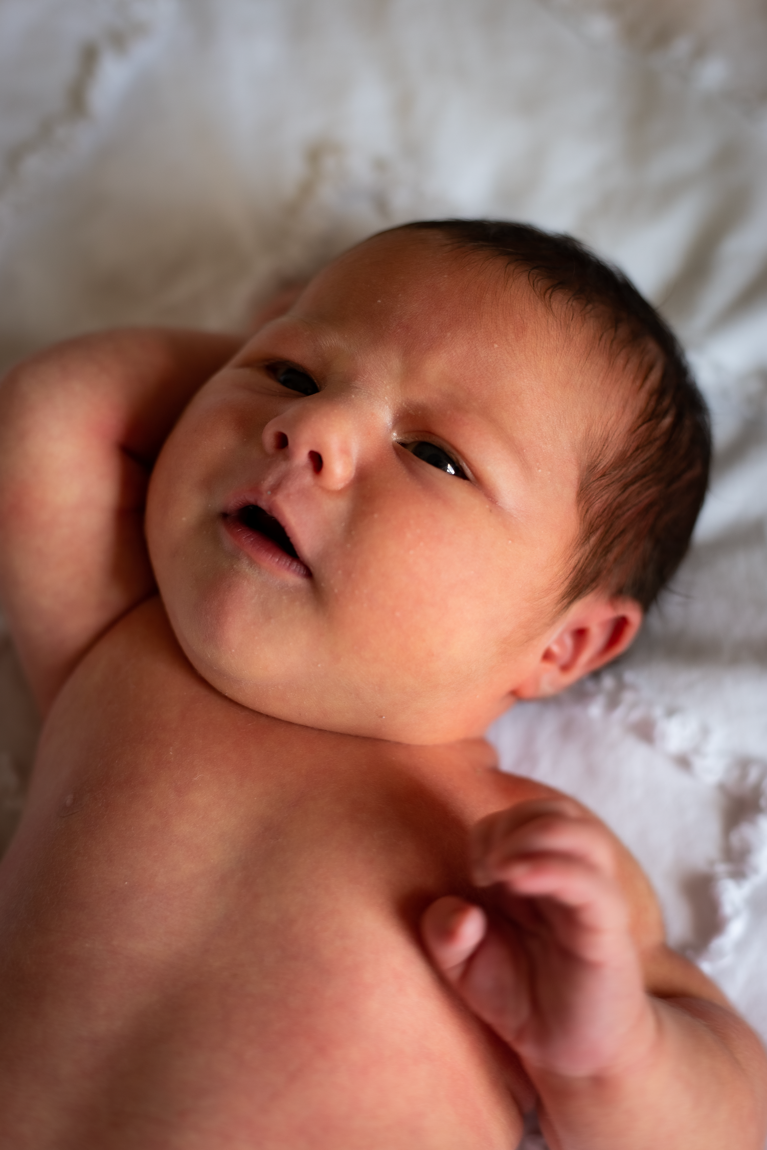 A close-up portrait of a newborn baby lying on a white blanket with eyes slightly open.