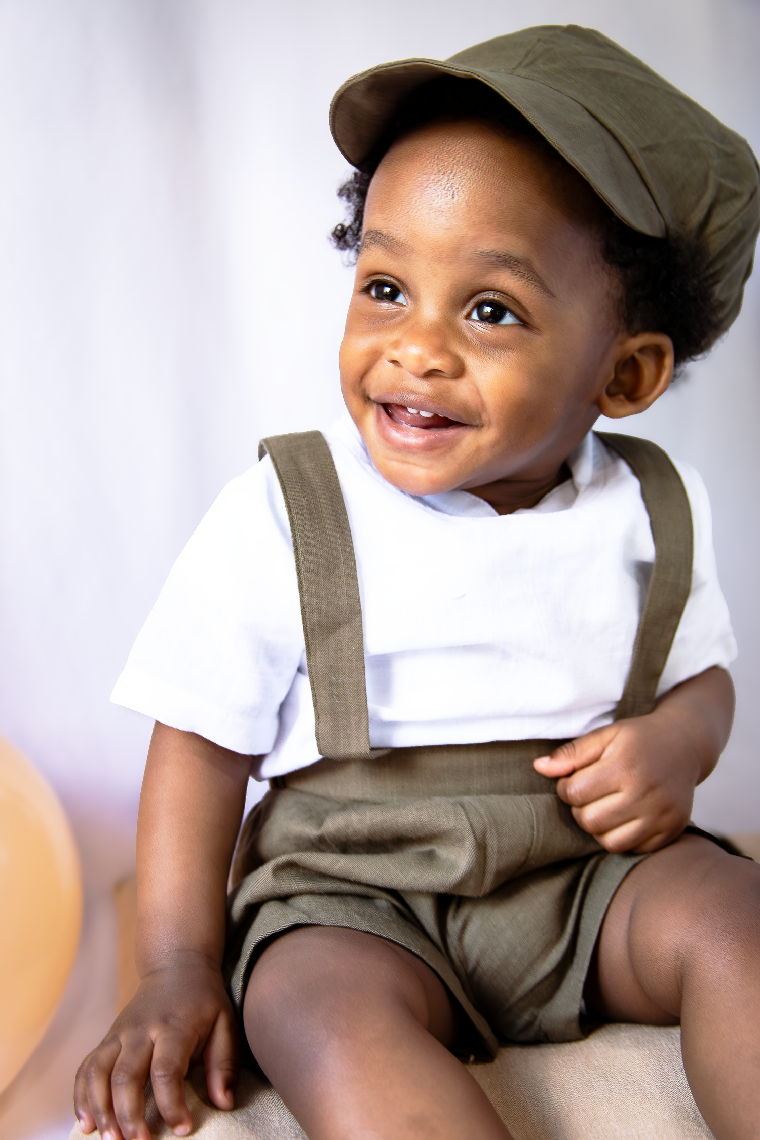Smiling toddler boy in olive green suspender shorts and matching cap sitting against light background.