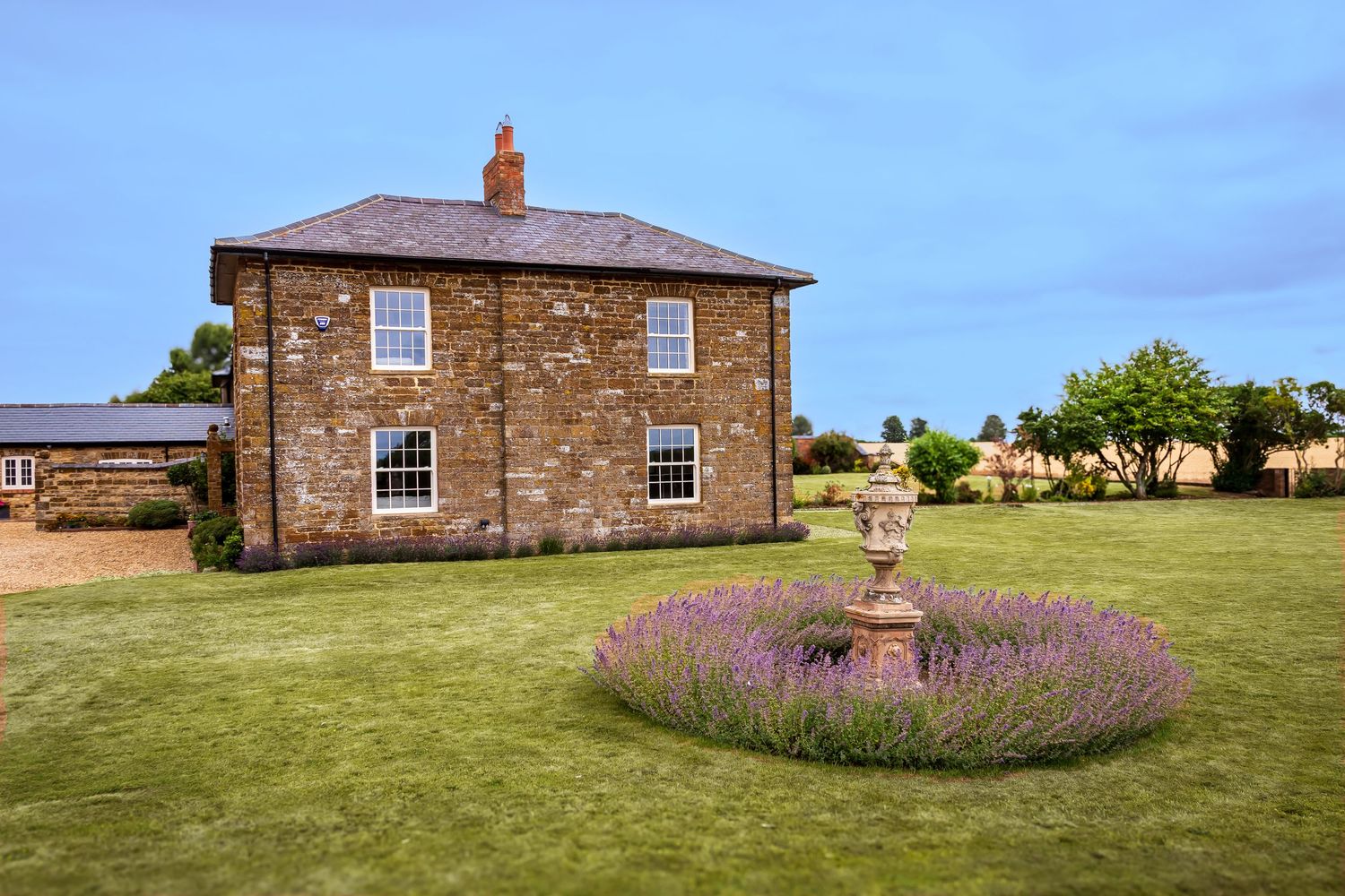 A charming stone country house with lawn and a circular lavender garden feature under blue sky.