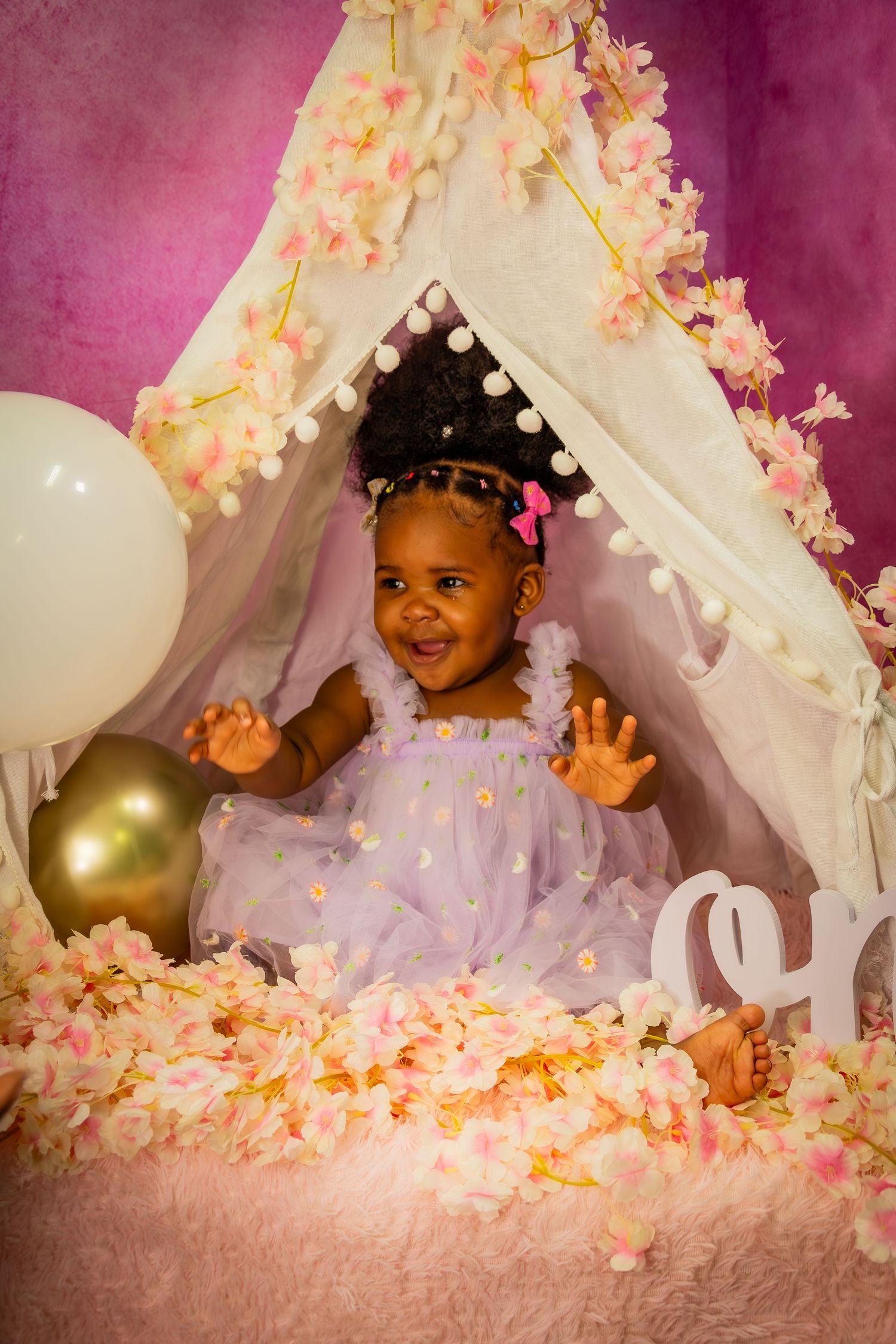 Happy baby girl in purple tutu dress laughing inside a floral canopy with balloons and flower petals.