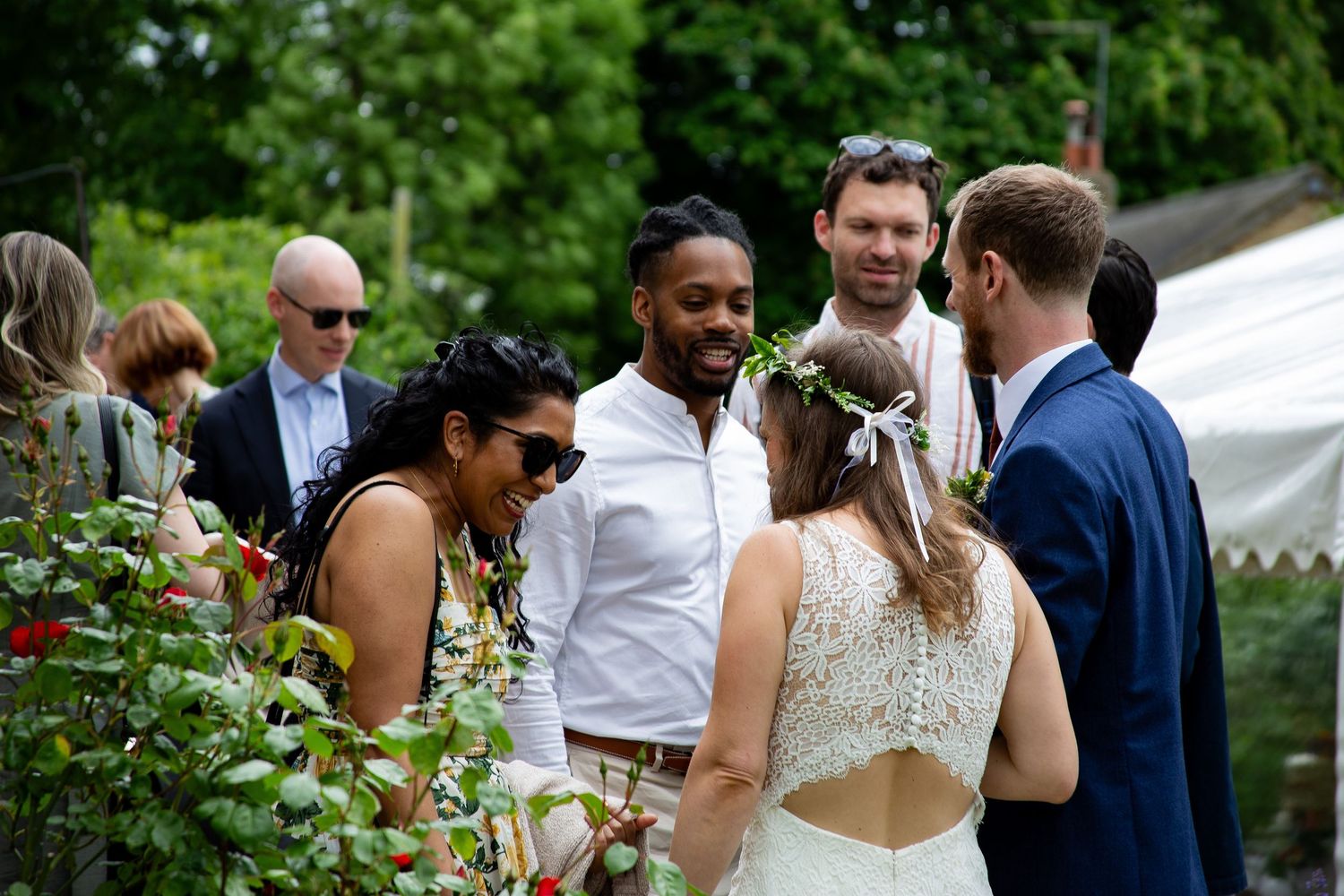 Wedding guests mingling outdoors in garden setting, bride in white floral crown chatting with guests near greenery.