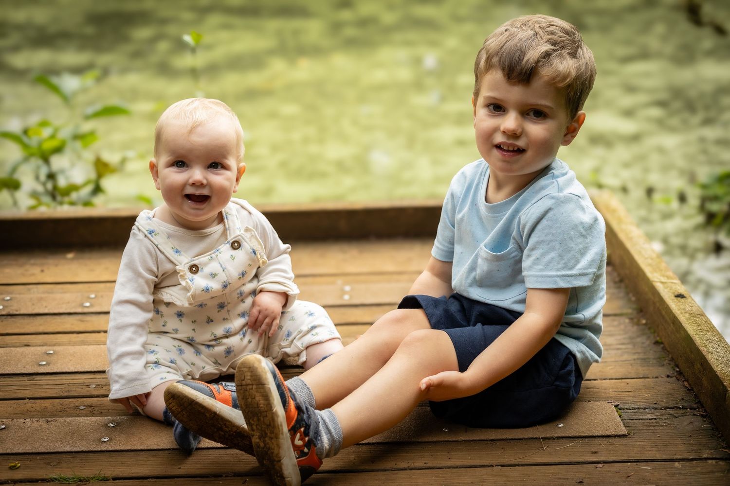 Two young brothers, a toddler and preschooler, sitting together on a wooden deck outdoors.