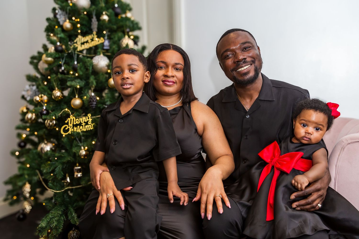 A joyful family of four in matching black outfits poses together near a Christmas tree.