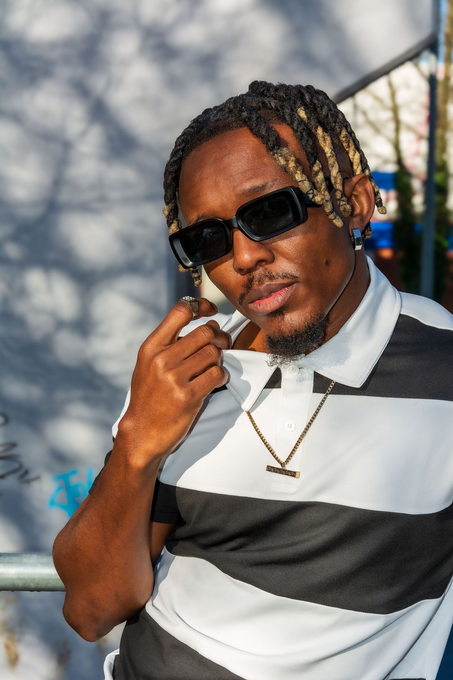 Stylish man modelling with braids and sunglasses wearing a striped polo and gold chain necklace outdoors.