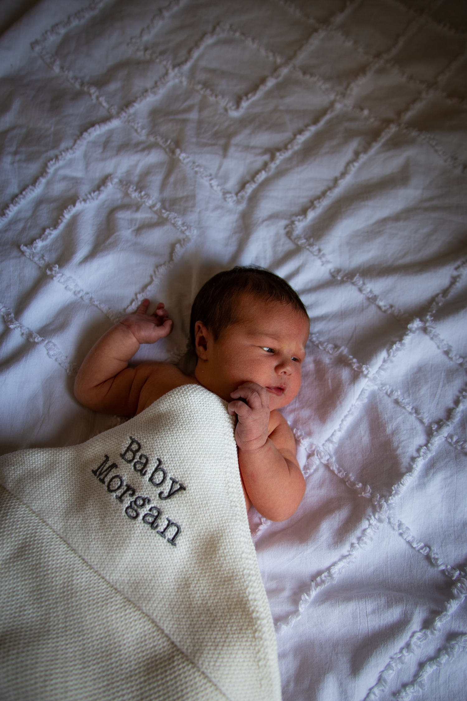 Newborn baby wrapped in personalized Baby Morgan cream knit blanket on white quilted bedding.