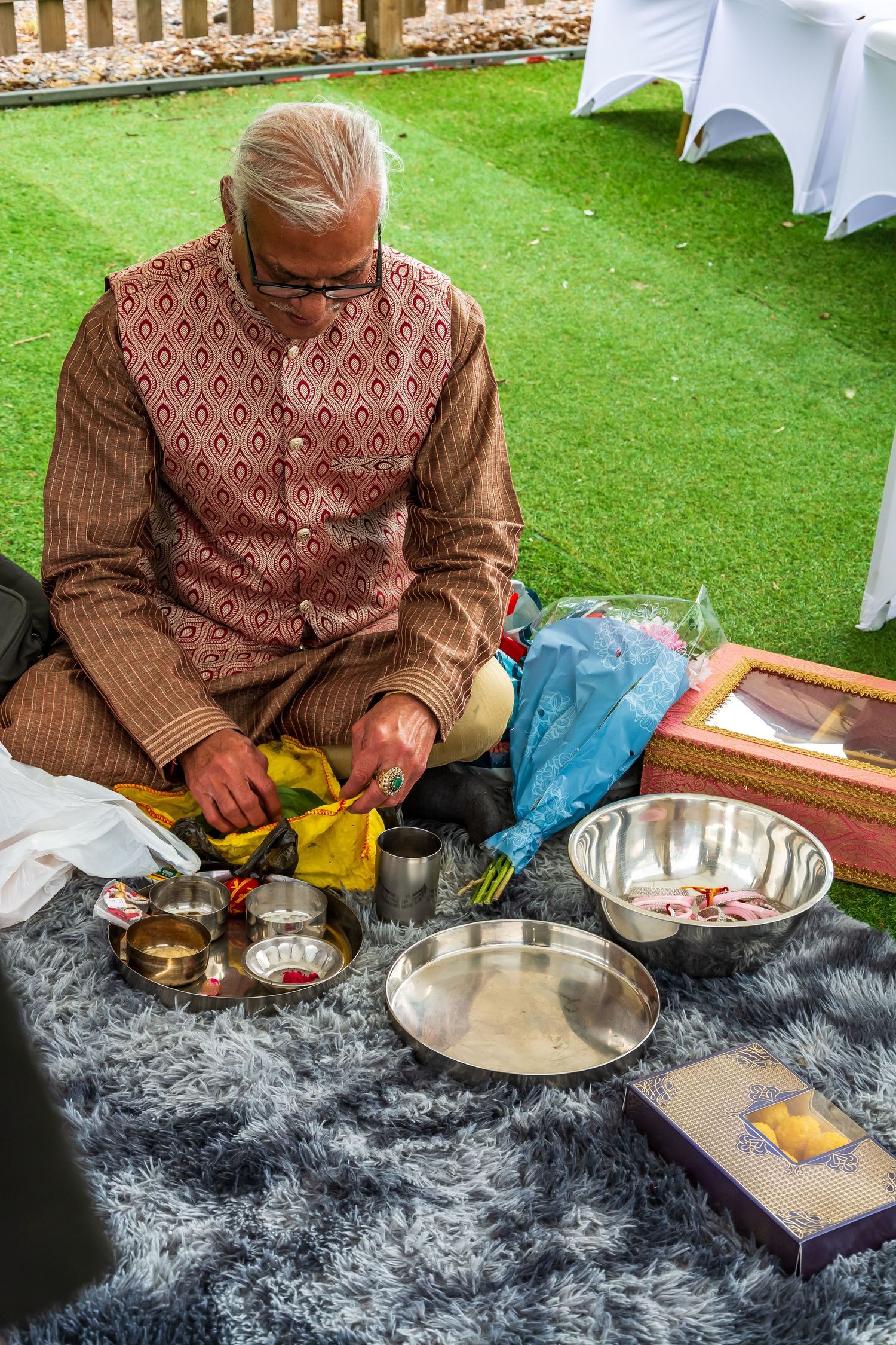 Elderly Indian man in traditional kurta preparing puja items on a rug outdoors.