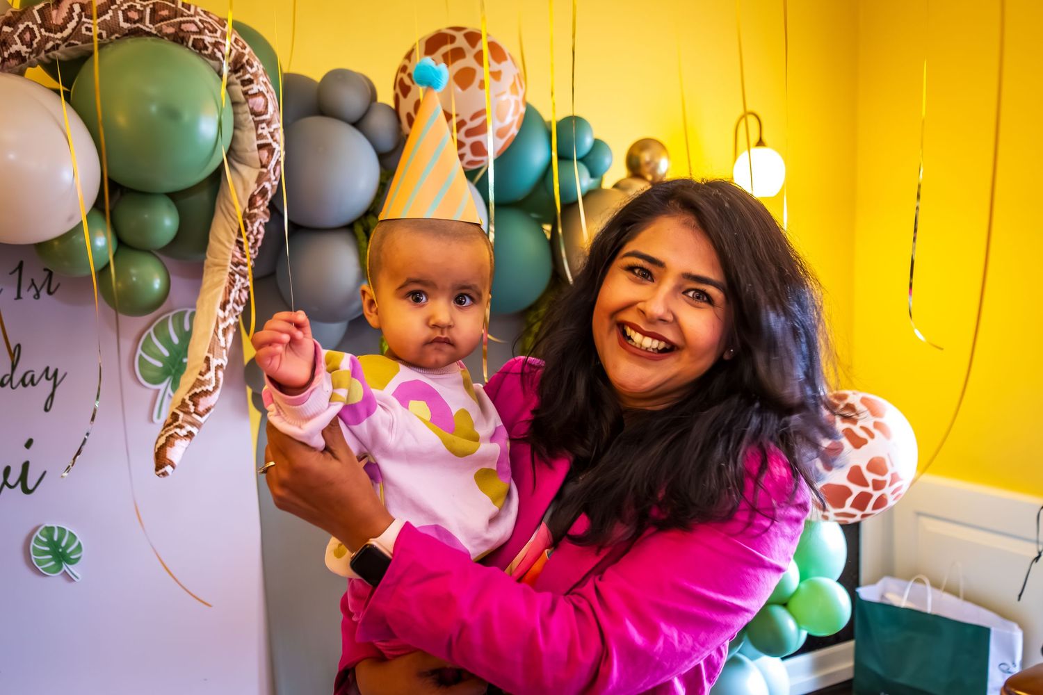 Smiling woman in pink holding baby with birthday hat at safari-themed first birthday party.