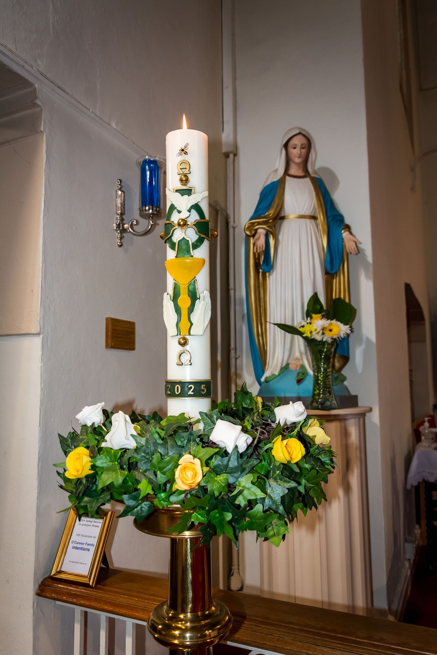Lit Easter paschal candle with white and yellow roses wreath beside a Virgin Mary statue in a church.