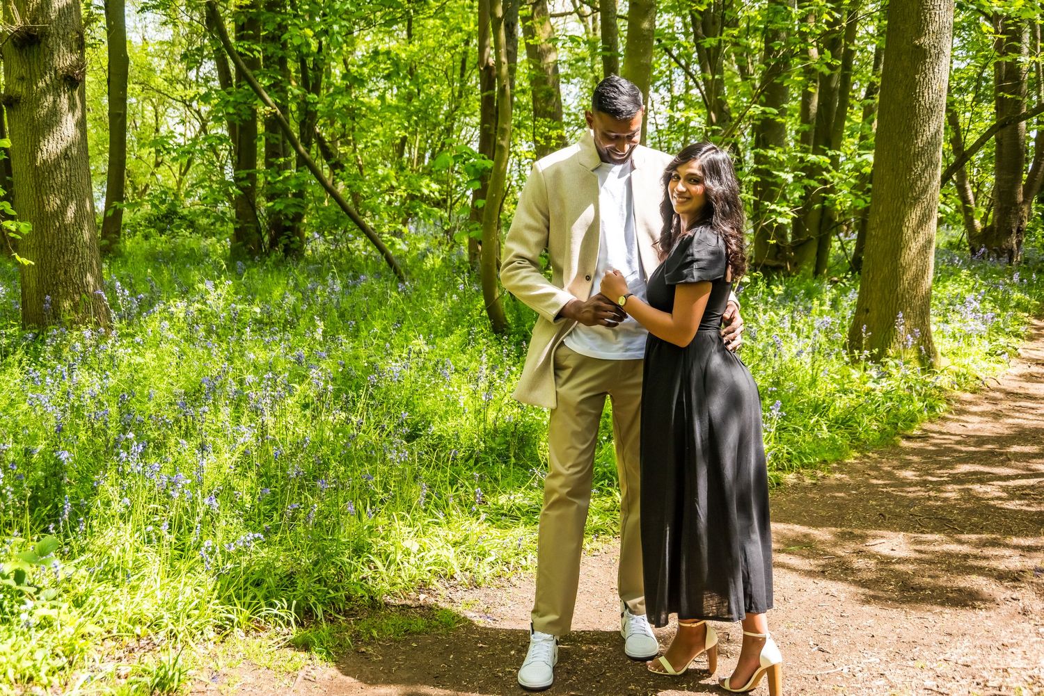 Couple in elegant attire walking on a woodland path surrounded by bluebells and lush green trees.