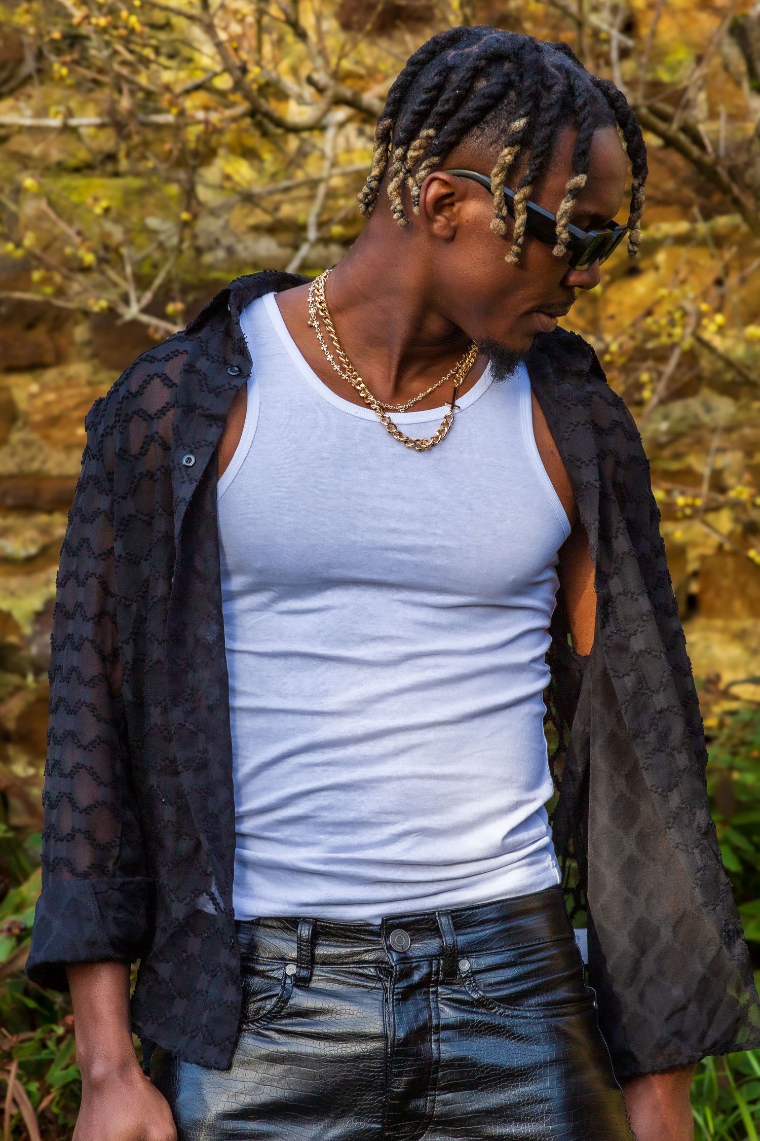 Athletic man modelling in white tank top and open black shirt wearing gold chains against a stone wall.