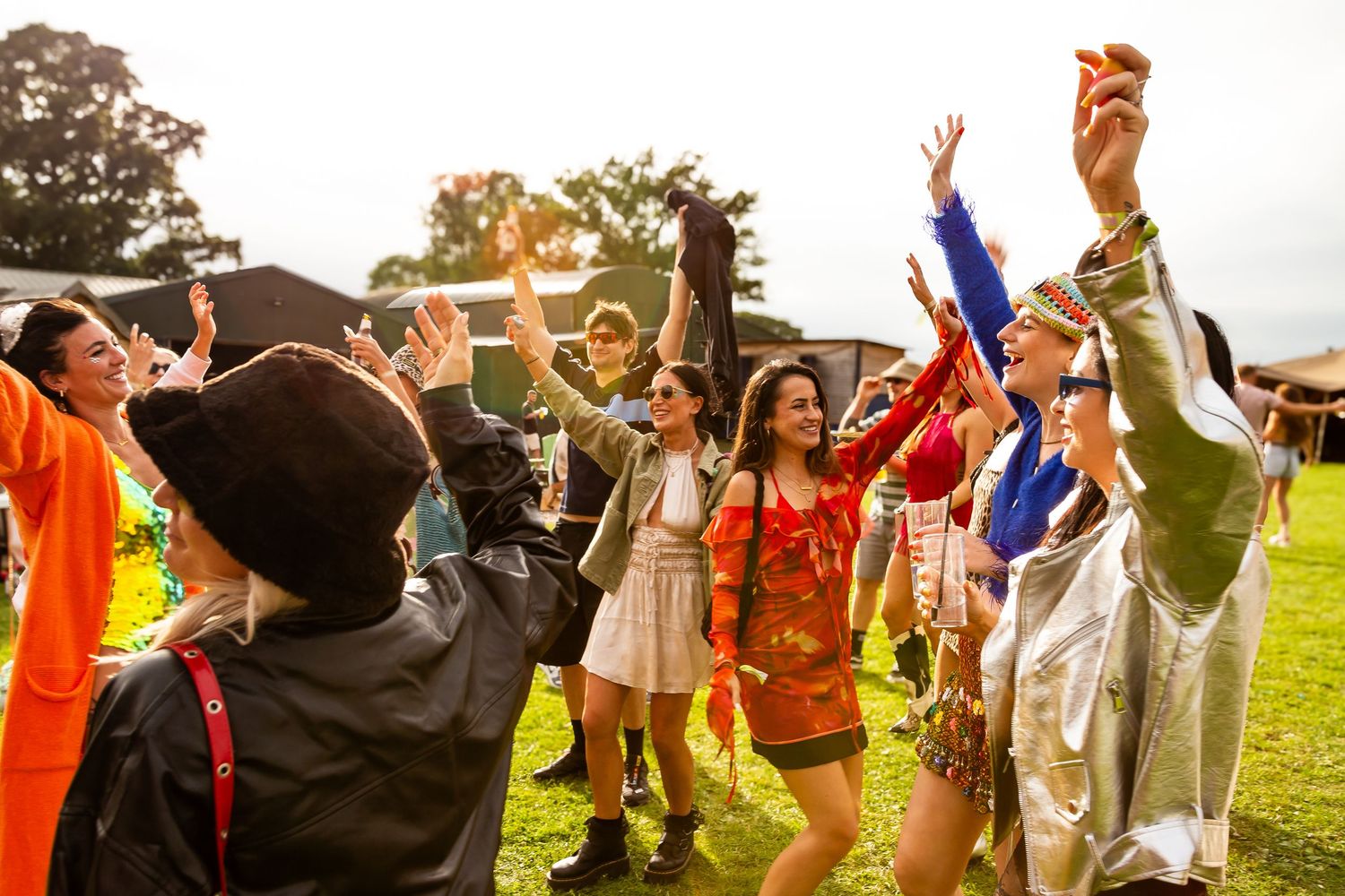 Group of colorfully dressed festival-goers dancing and celebrating with raised arms at an outdoor music festival.