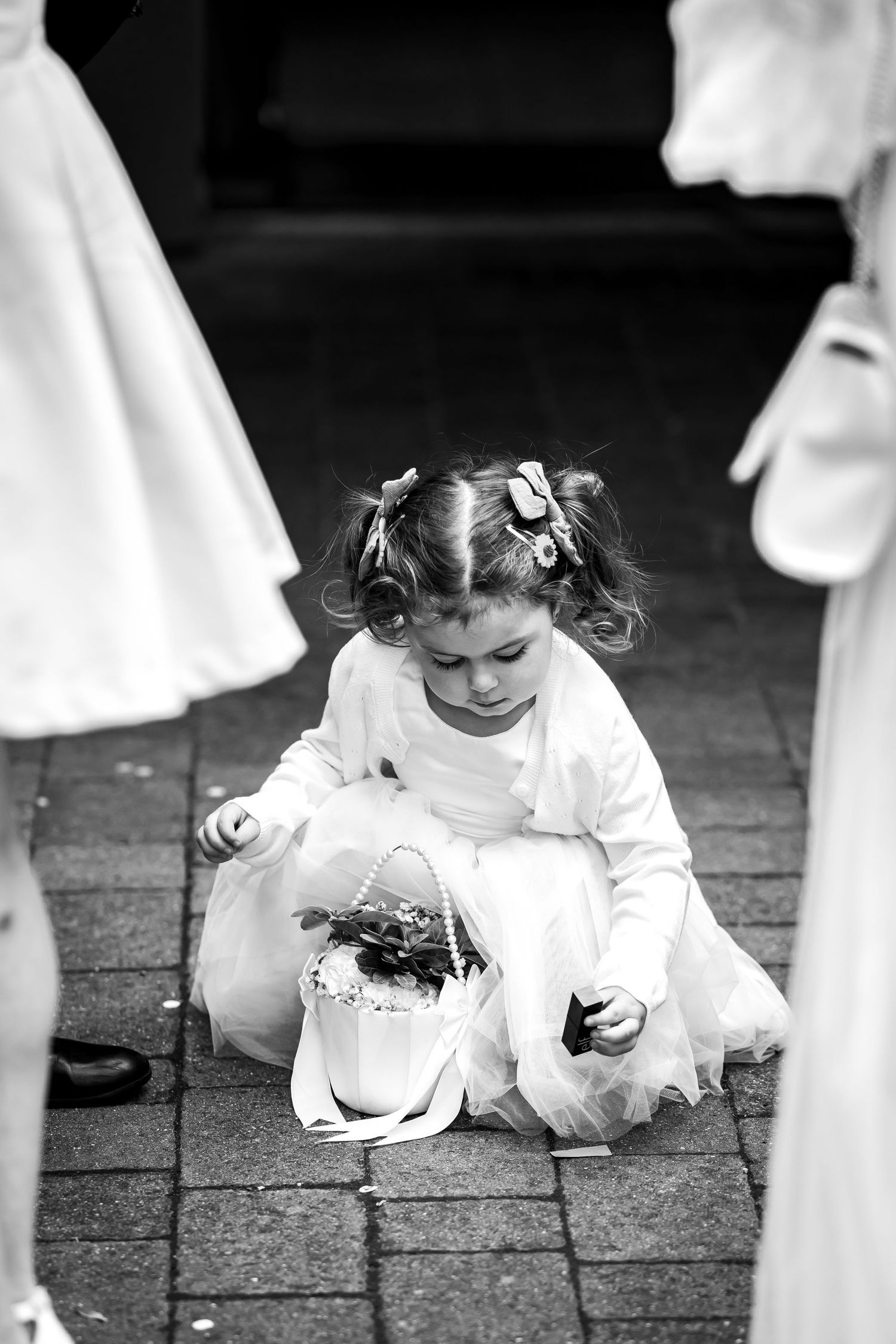 Black and white photo of young flower girl in white dress crouching on cobblestone ground with flower basket.