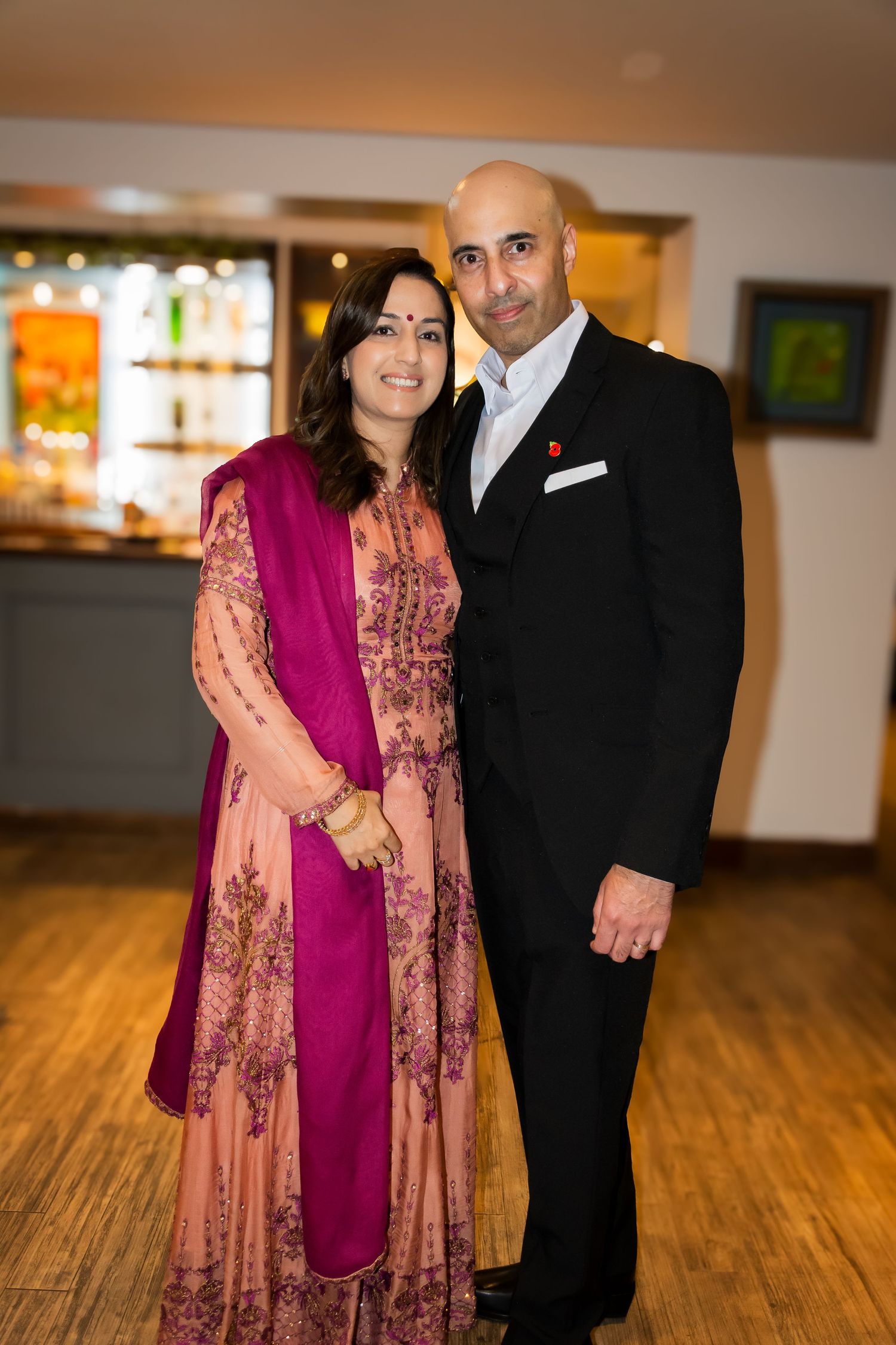 South Asian couple posing together, woman in pink embroidered salwar kameez and man in black suit.