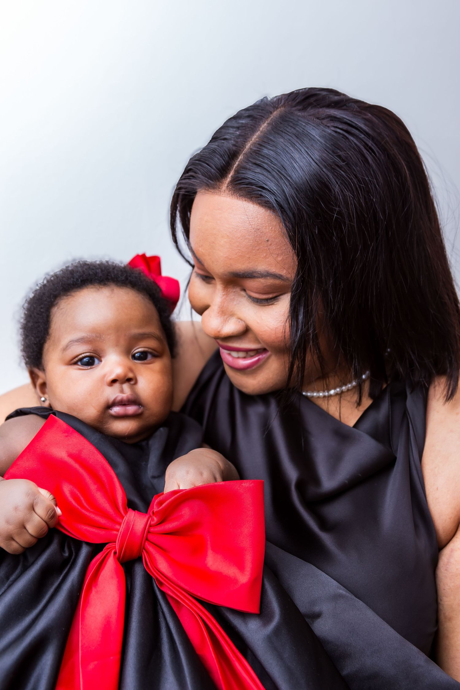A mother in a black dress smiles warmly at her baby girl wearing a red bow dress.