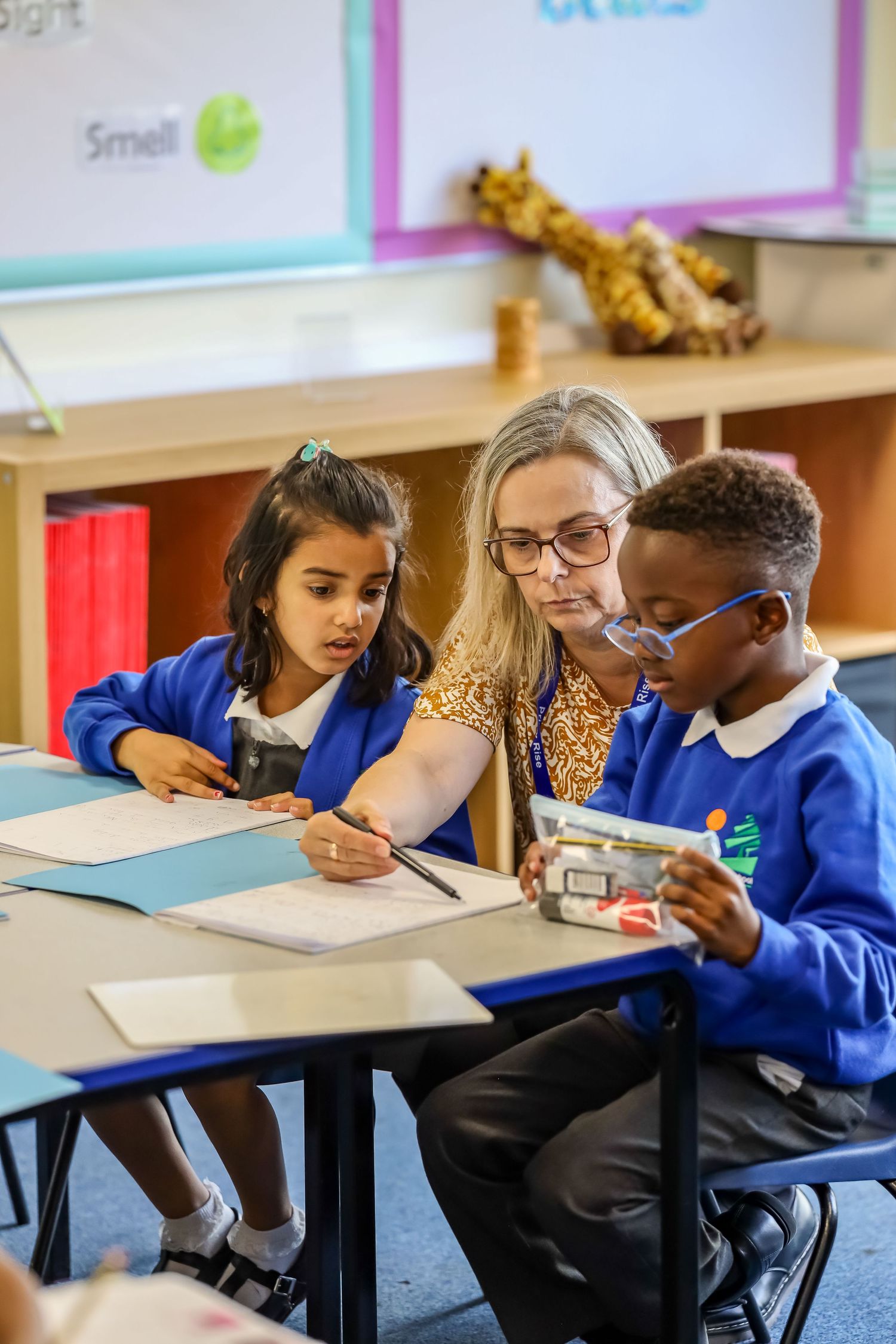 Teacher helping two young students with schoolwork at a classroom desk.