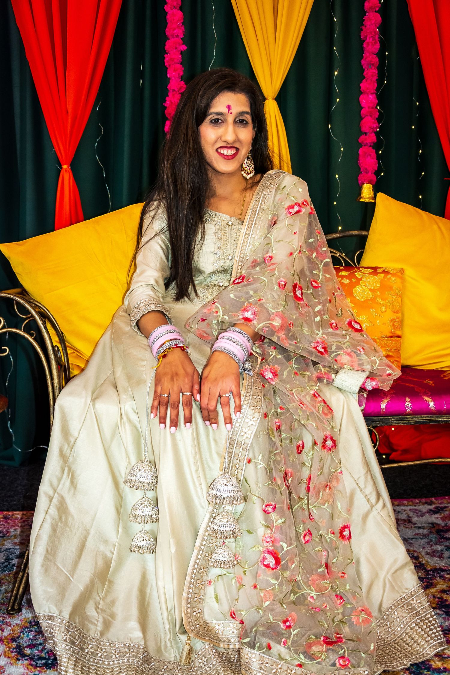 Young Indian woman in elegant cream lehenga with floral dupatta seated at a Mehndi ceremony.