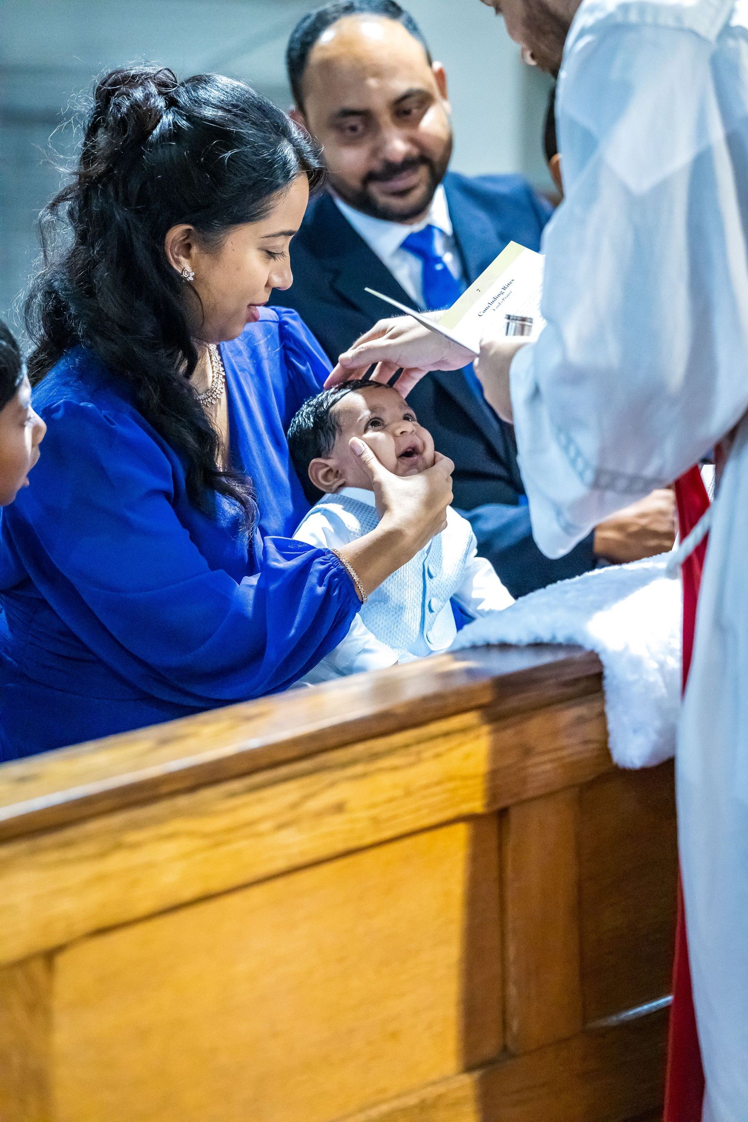 Priest performing infant baptism with water as mother in blue dress holds baby at the font.