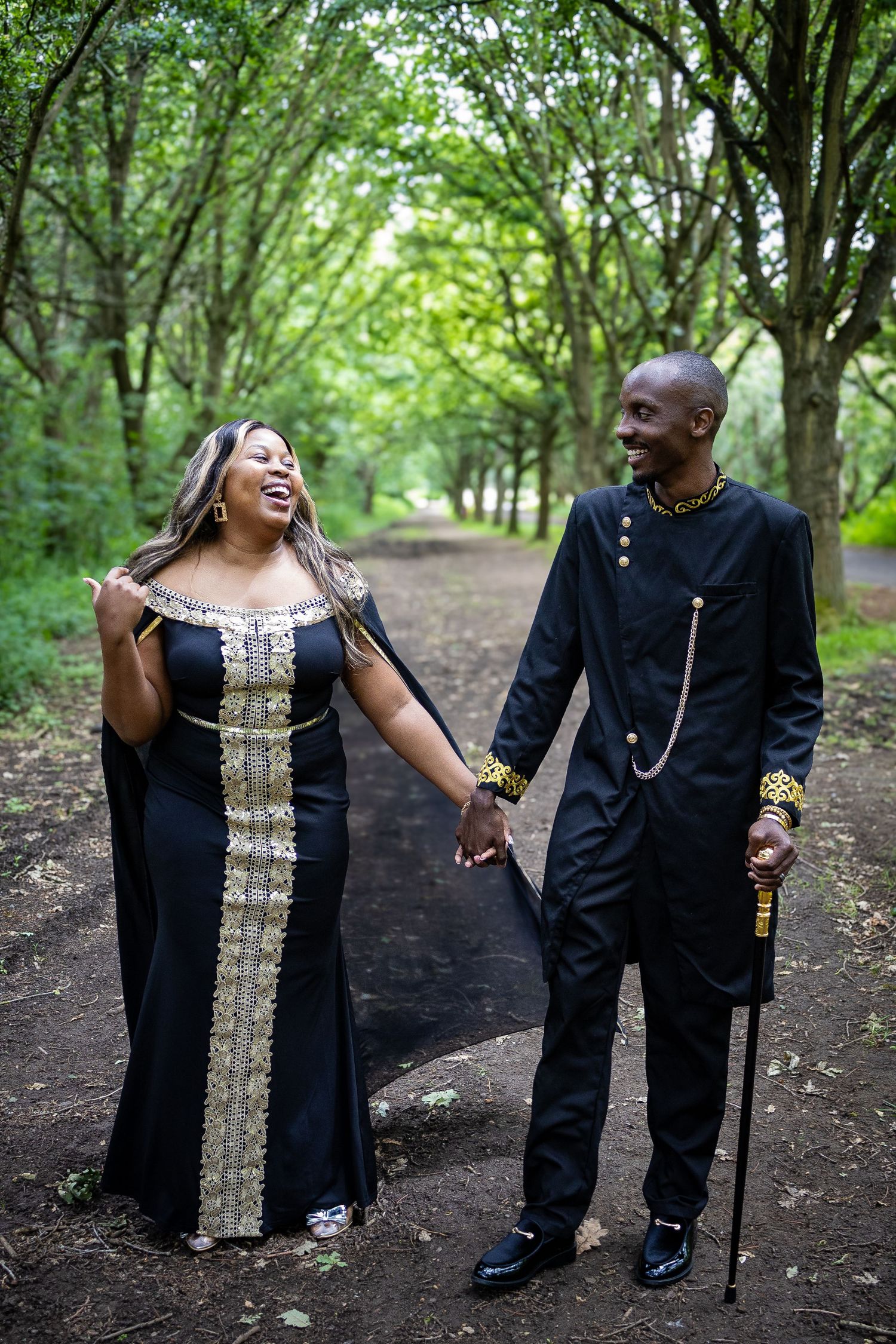 Couple in elegant black traditional African attire holding hands and smiling on a tree-lined path.