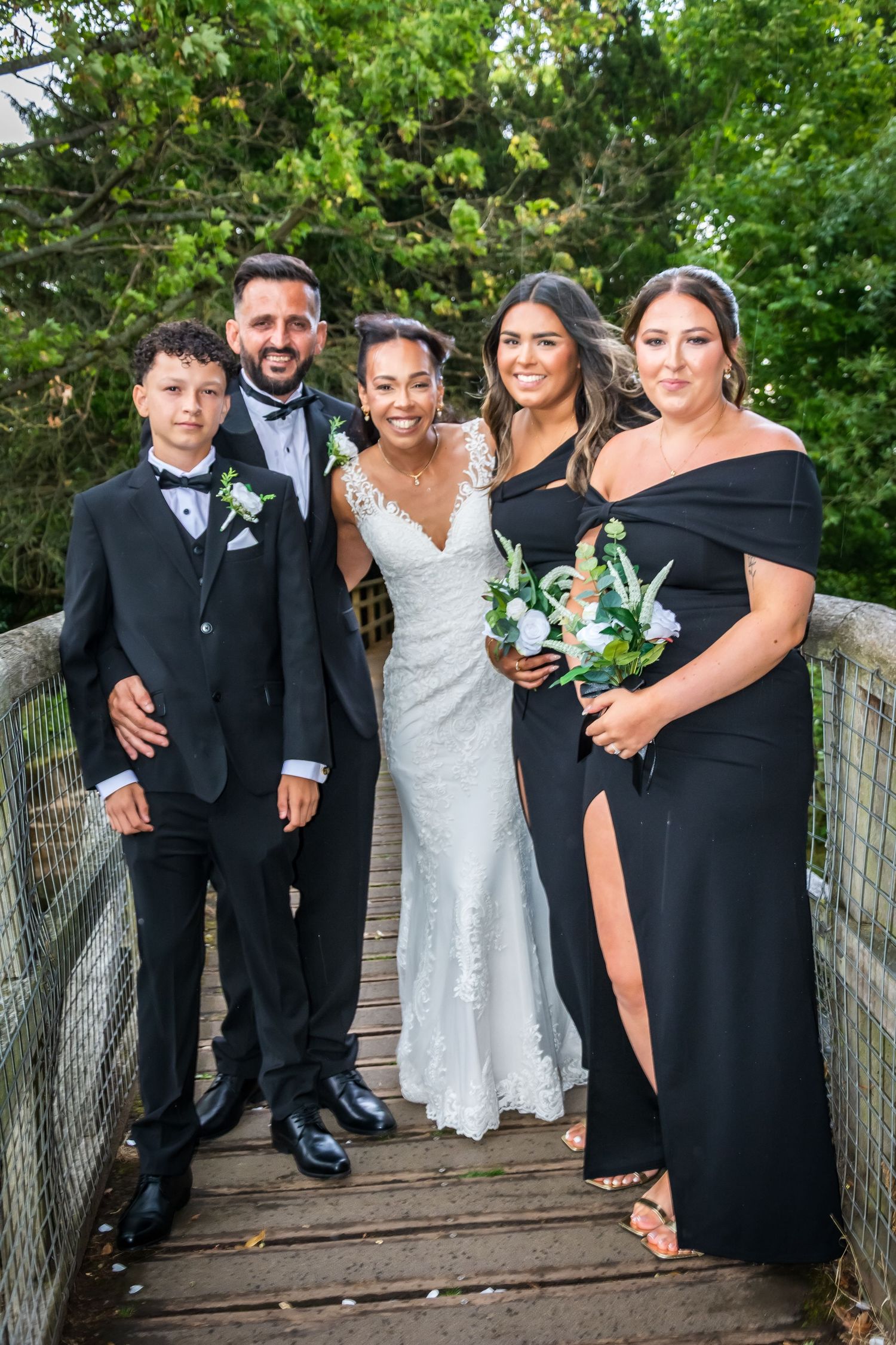 Wedding party posing on wooden bridge, bride in lace gown with groom in tuxedo, two bridesmaids in black dresses and young boy.