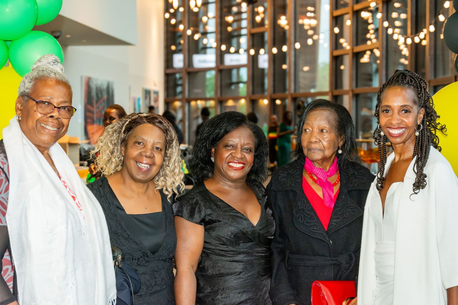 Five women smiling together at a celebration with green and yellow balloon decorations.