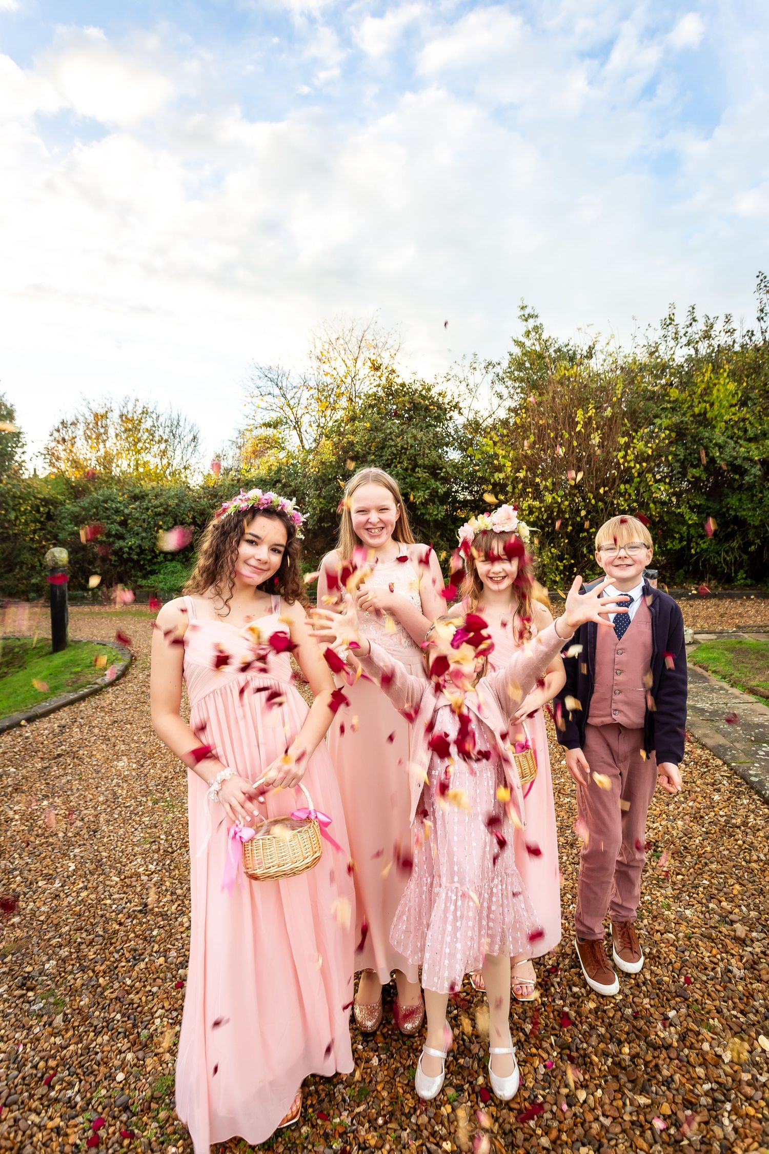 Four wedding party members in pink dresses tossing rose petals in a garden path.
