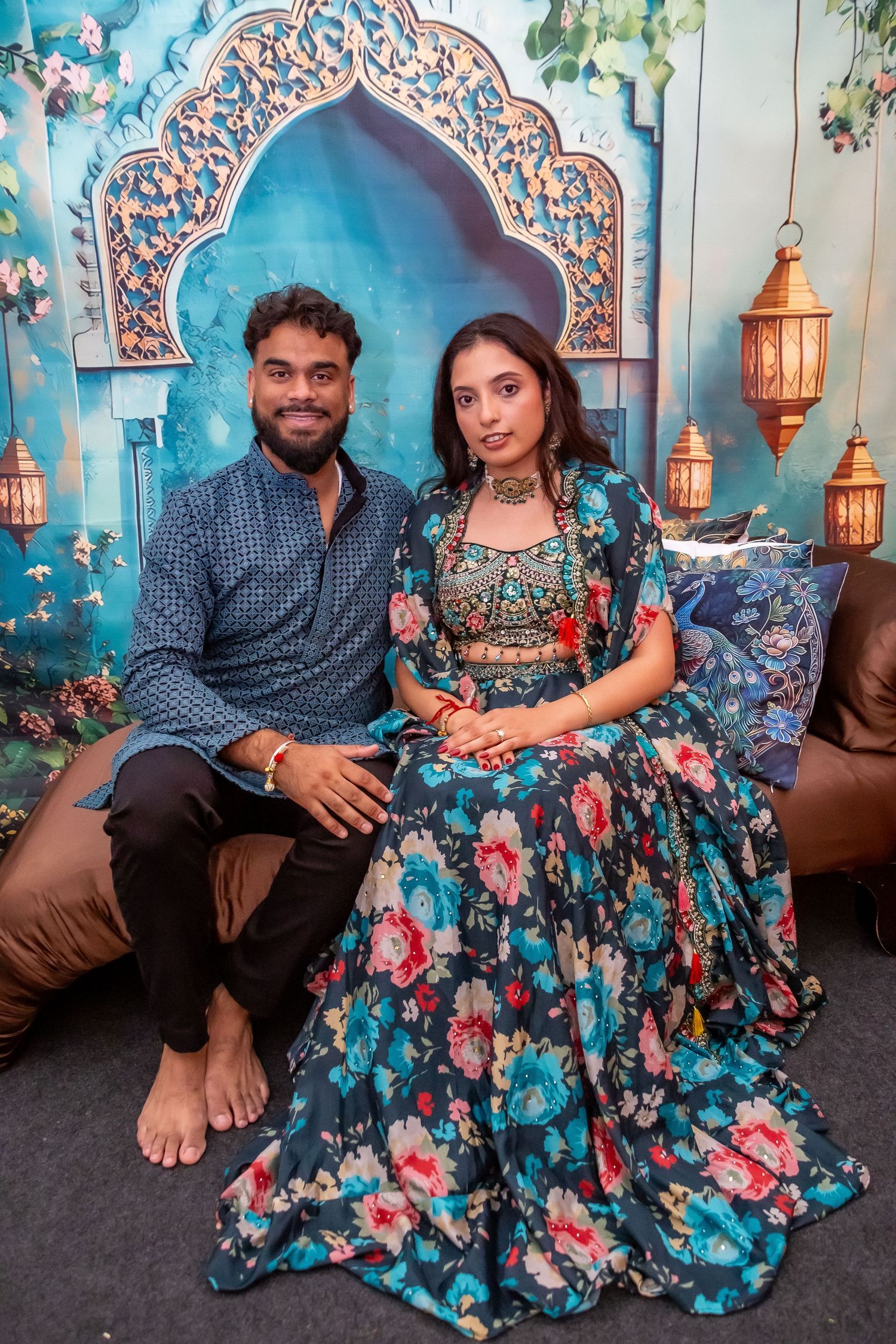 Indian couple sitting together in traditional floral outfits against decorative Mughal arch backdrop.