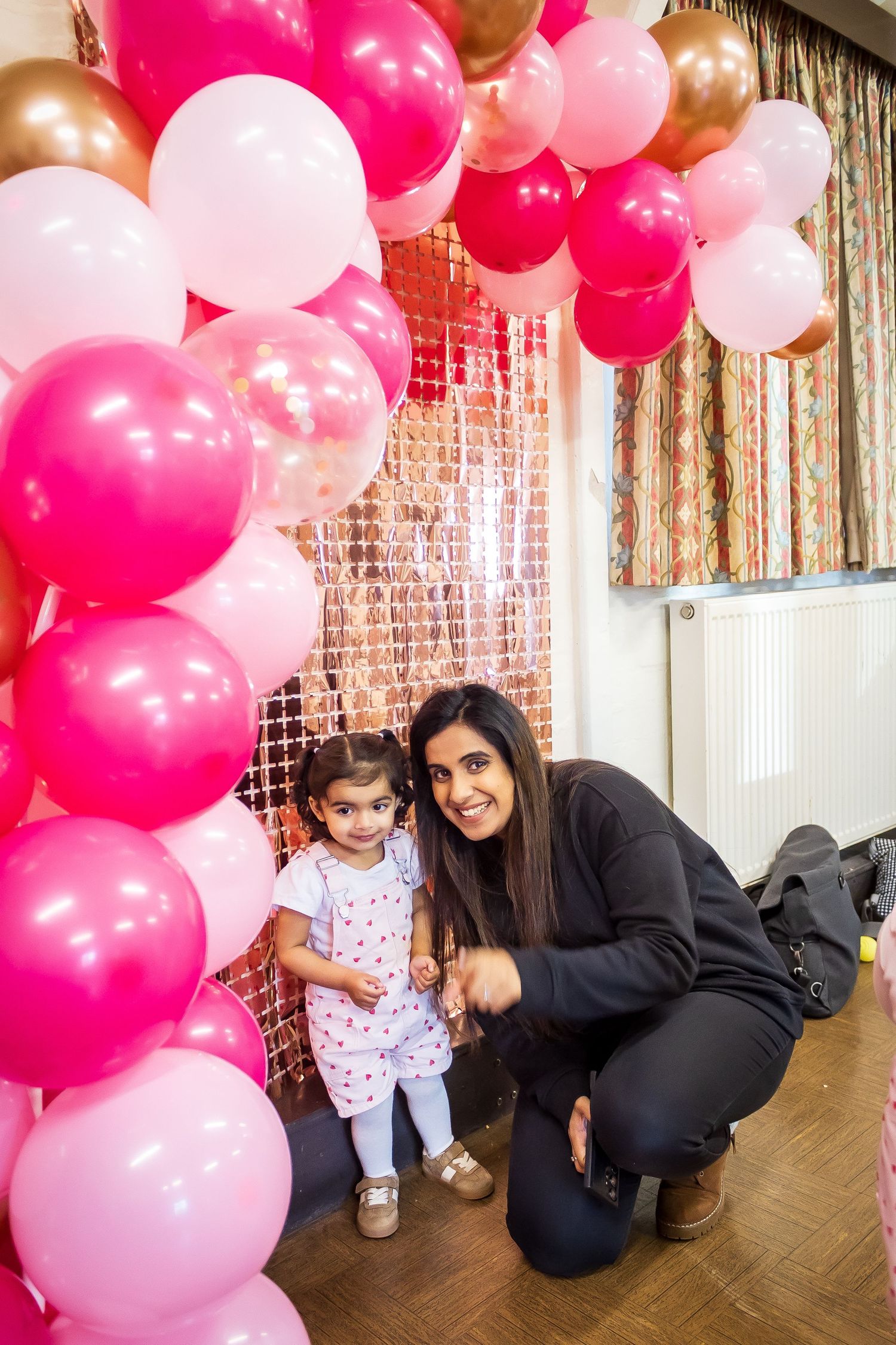 Woman crouching beside young girl under a pink and gold balloon arch at a birthday party.