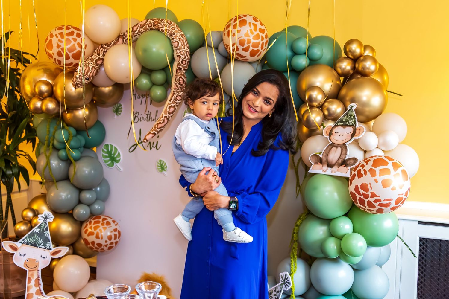 Woman in blue dress holding toddler in front of safari-themed balloon arch at birthday party.