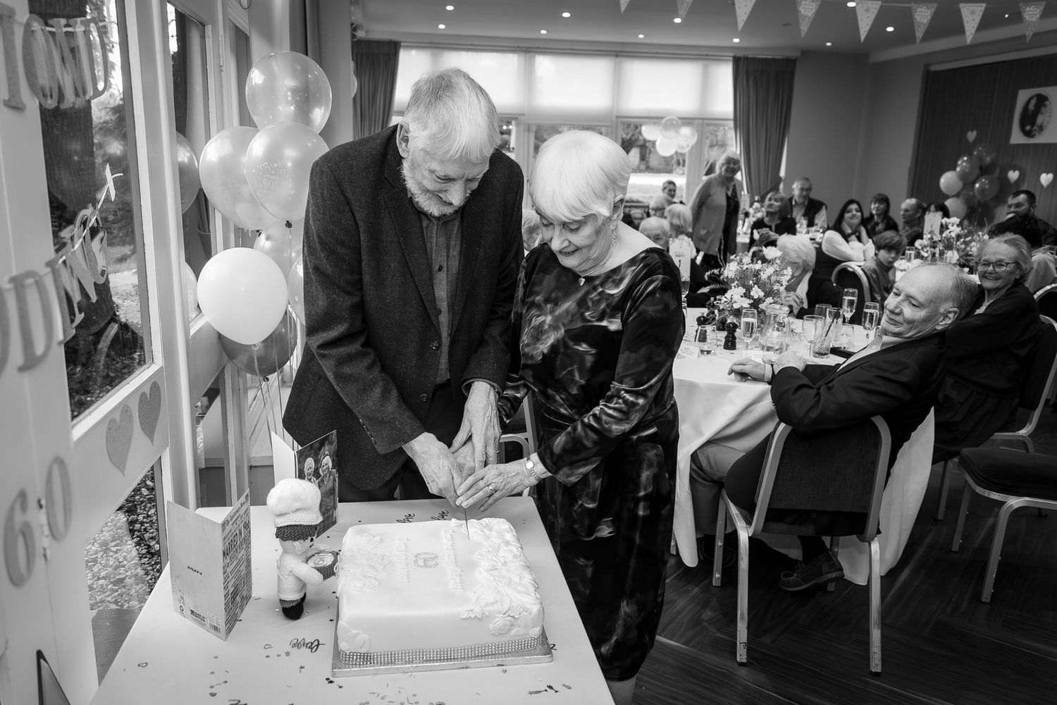Elderly couple cutting a white wedding anniversary cake at a celebration party with balloons in black and white photo.