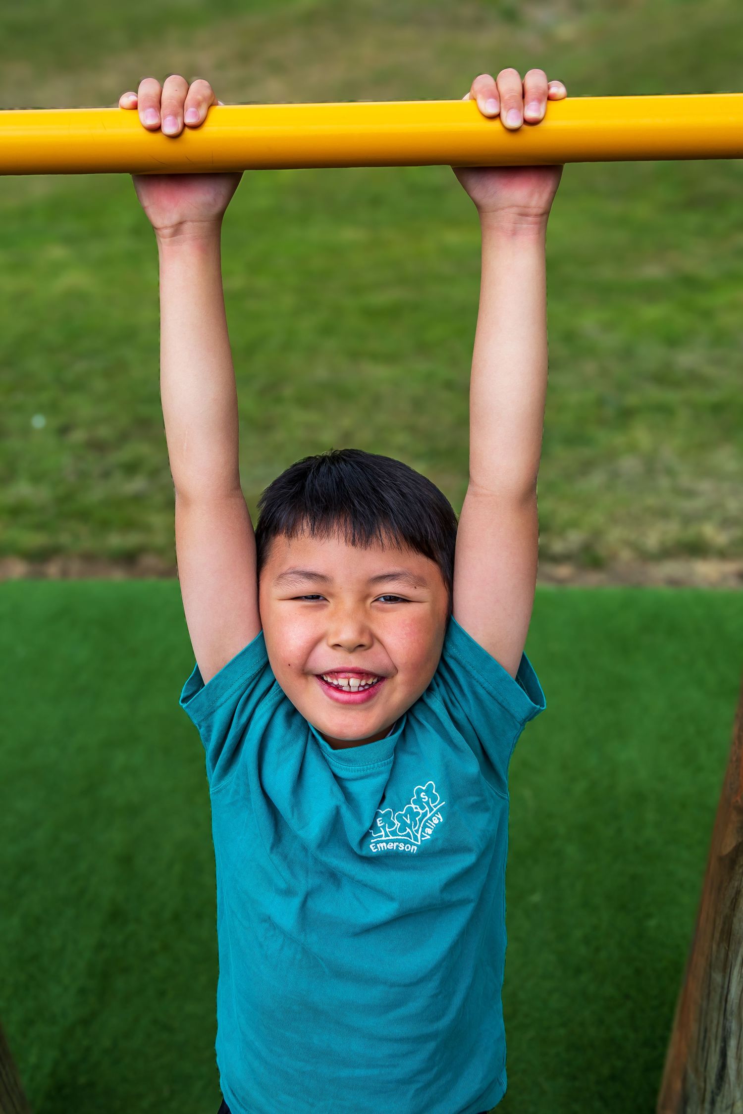 Happy young boy hanging from a yellow playground bar, smiling outdoors on green grass.